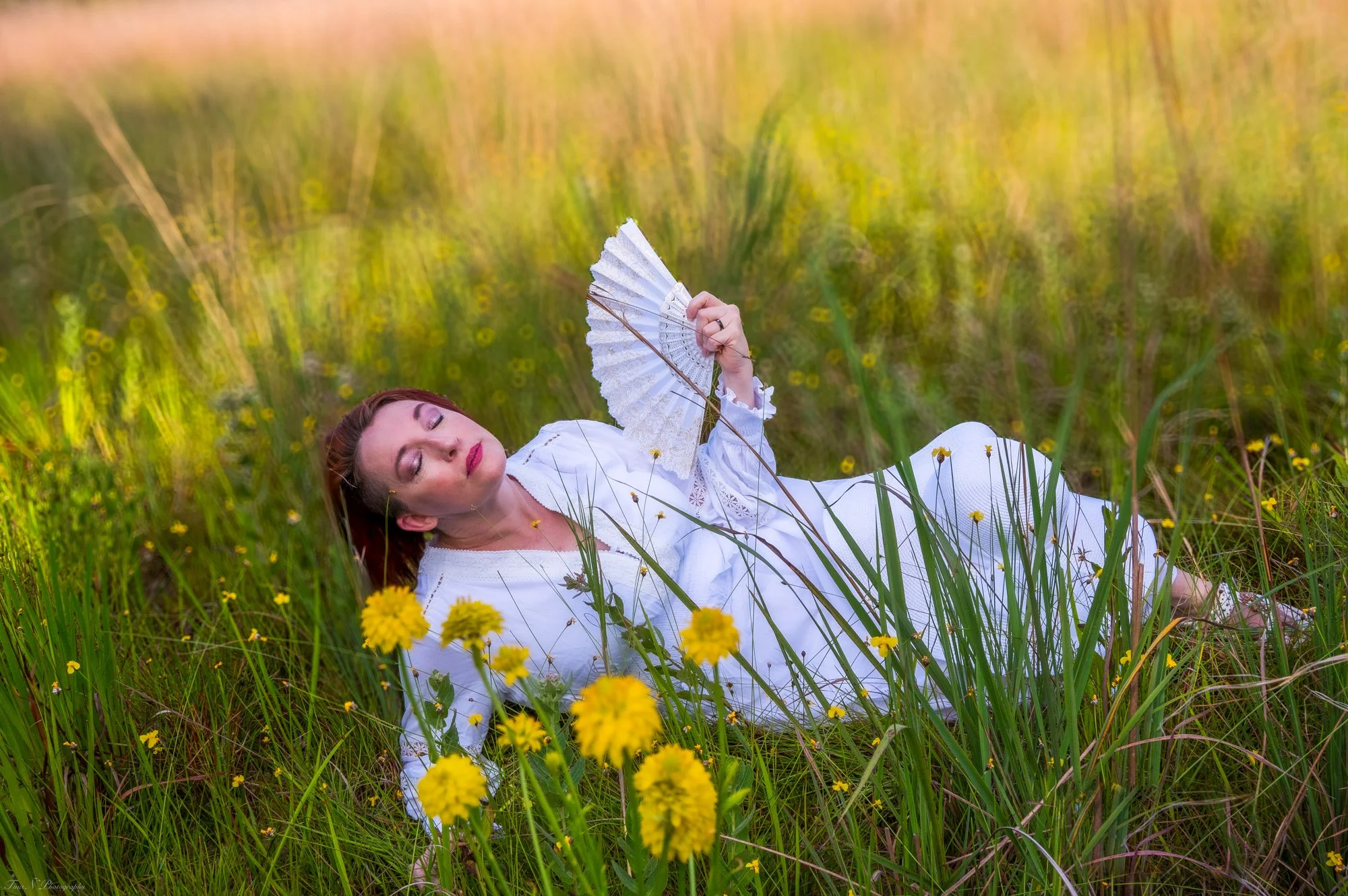 A woman in white lying in a field of yellow flowers holding a white lace fan.