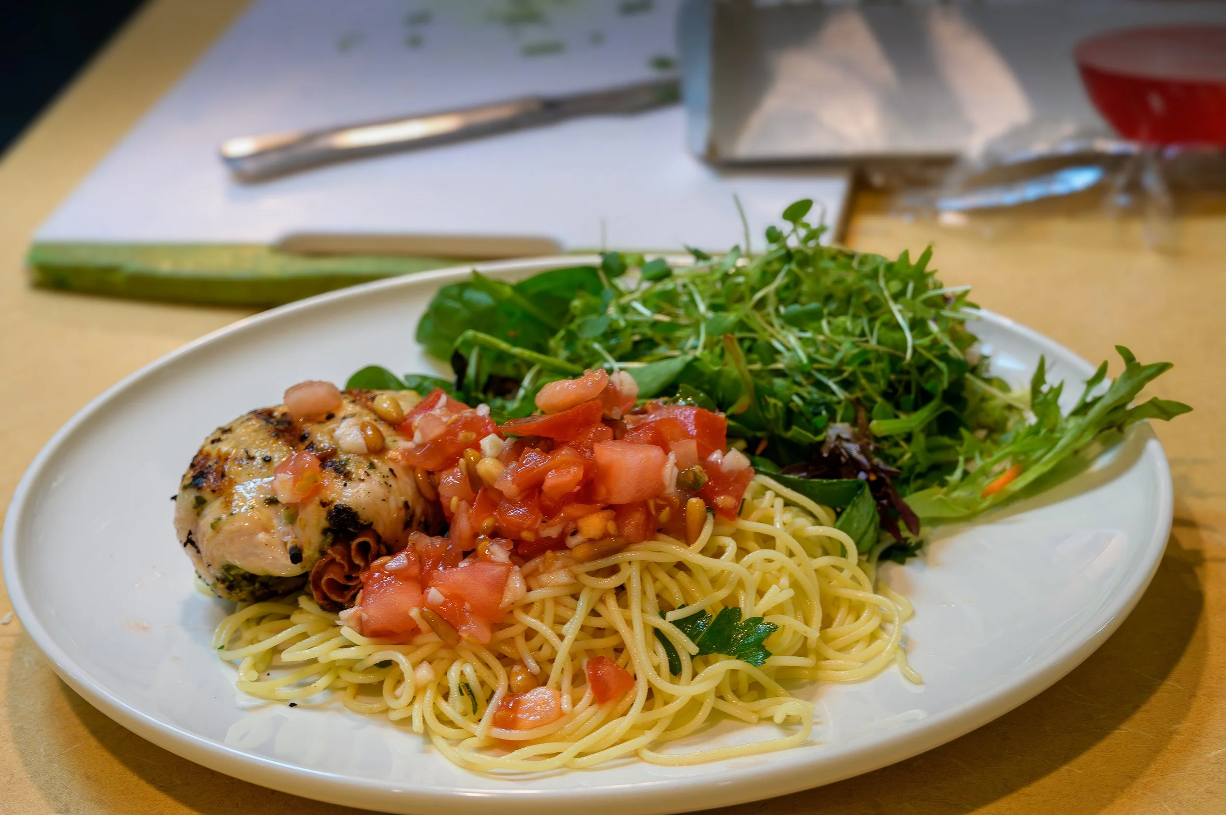 A white plate with grilled chicken topped with salsa, spaghetti, and a side salad of mixed greens. In the background, there are utensils and a napkin on a wooden table.