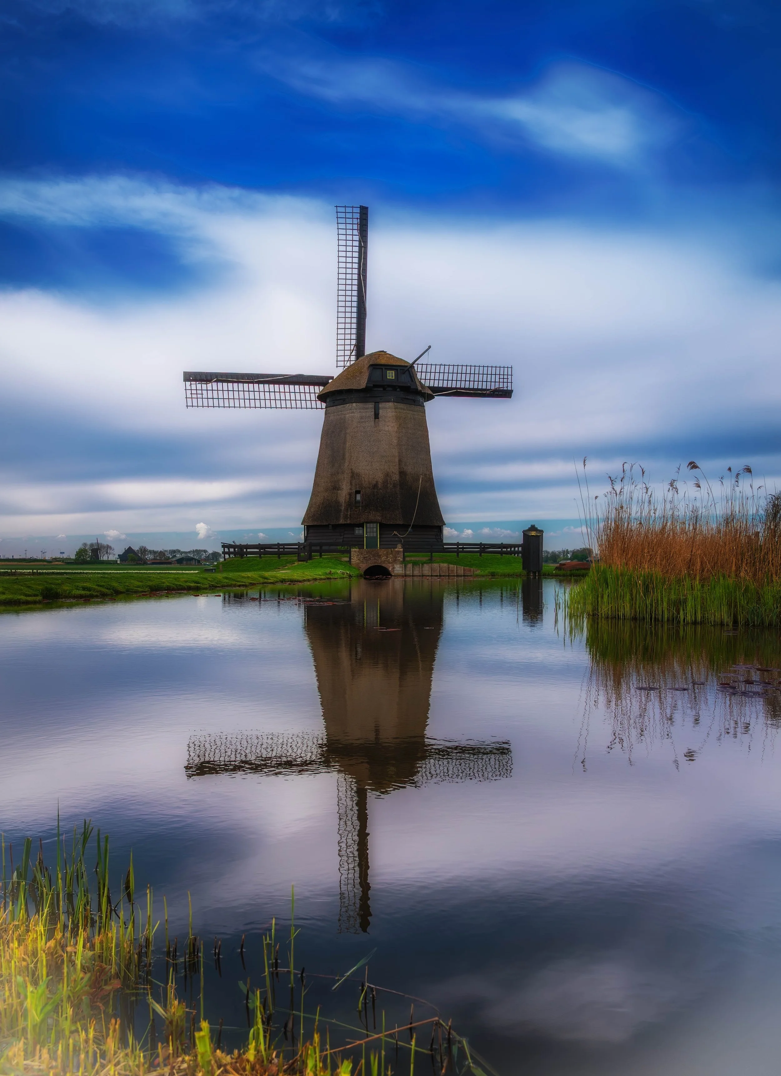 Hoorn, Amsterdam. A windmill beside a calm water canal, with a blue, cloudy sky and green landscape reflecting in the water.