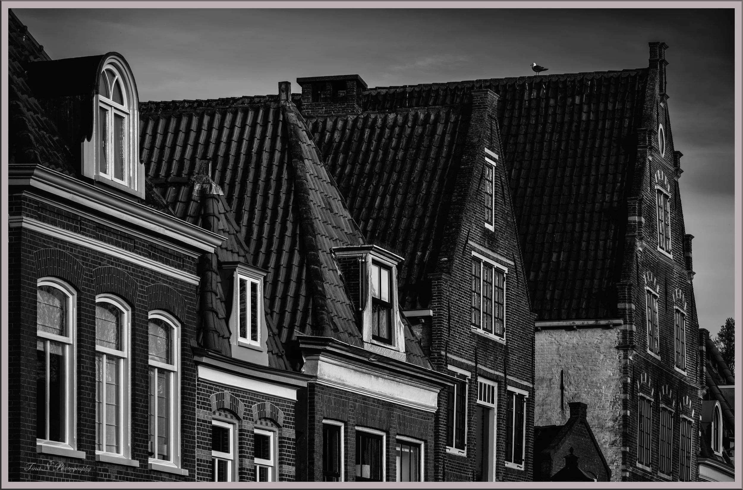 Black and white photo of old brick buildings with pointed roofs, dormer windows, and decorative brickwork.