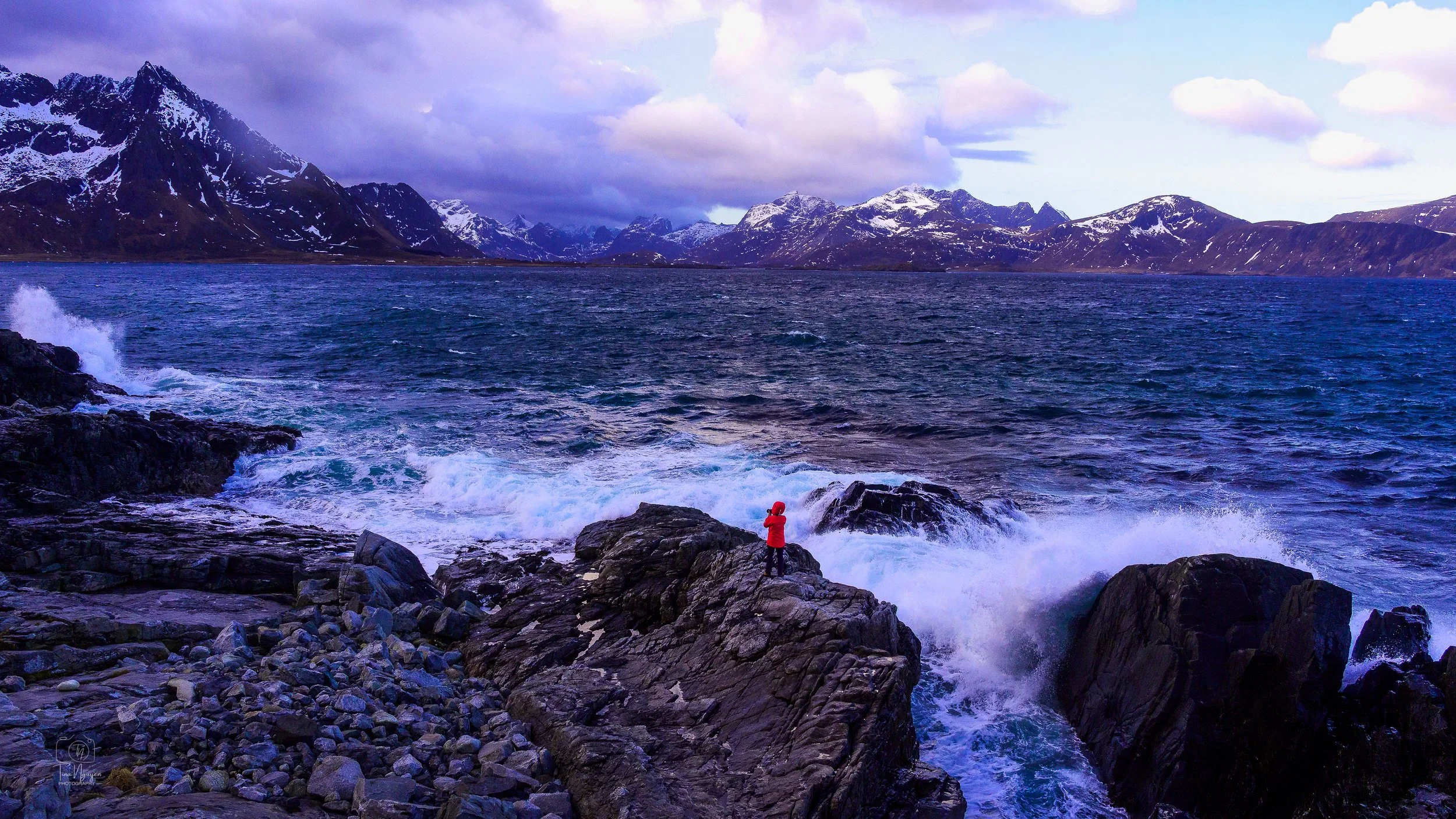 A person in a red jacket standing on rocks by the ocean with mountains in the background and a cloudy sky.