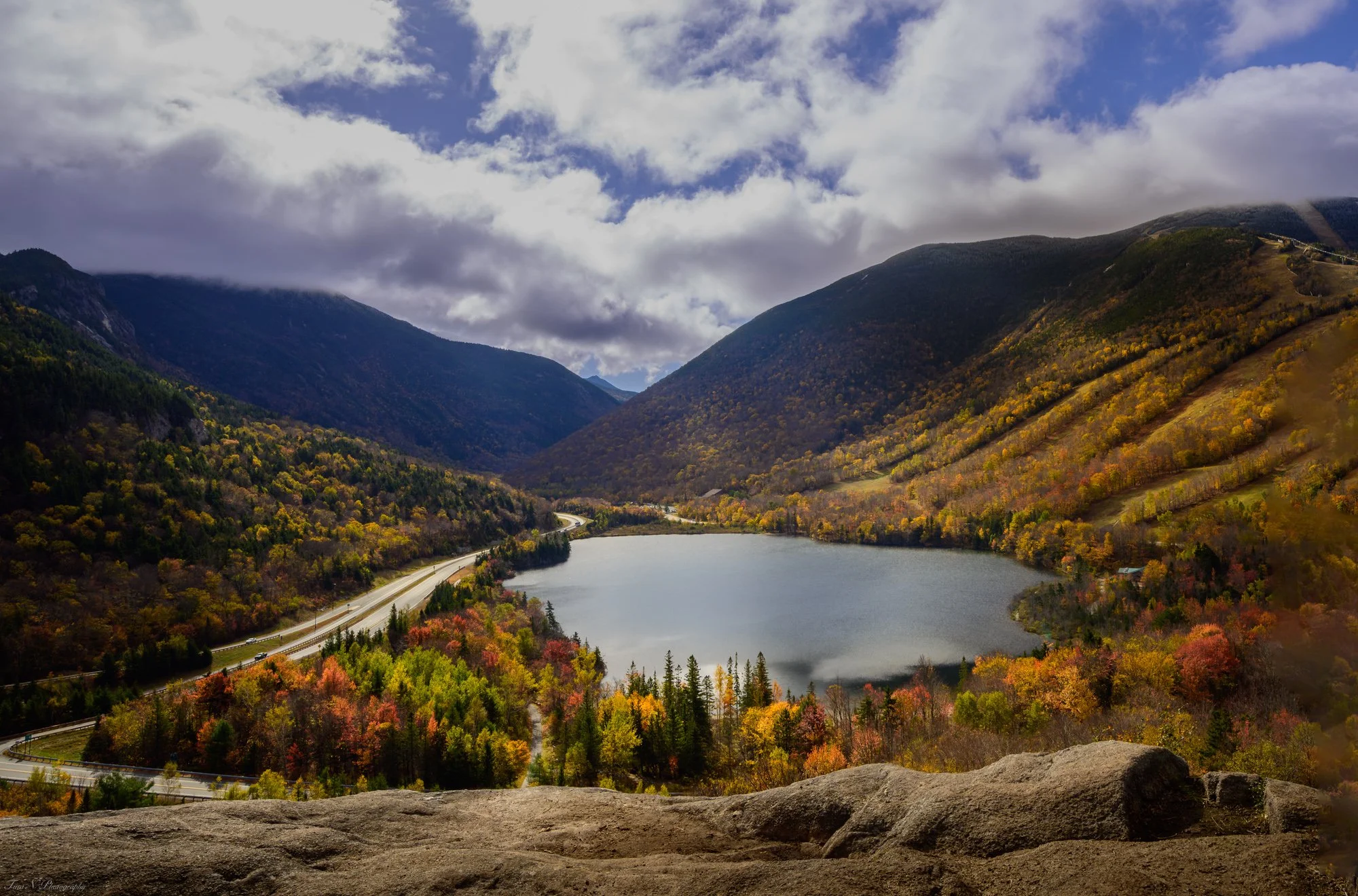 Echo Lake, NH. Autumn landscape of a mountain lake surrounded by colorful fall foliage, with mountains, a partly cloudy sky, and a winding road in the background.