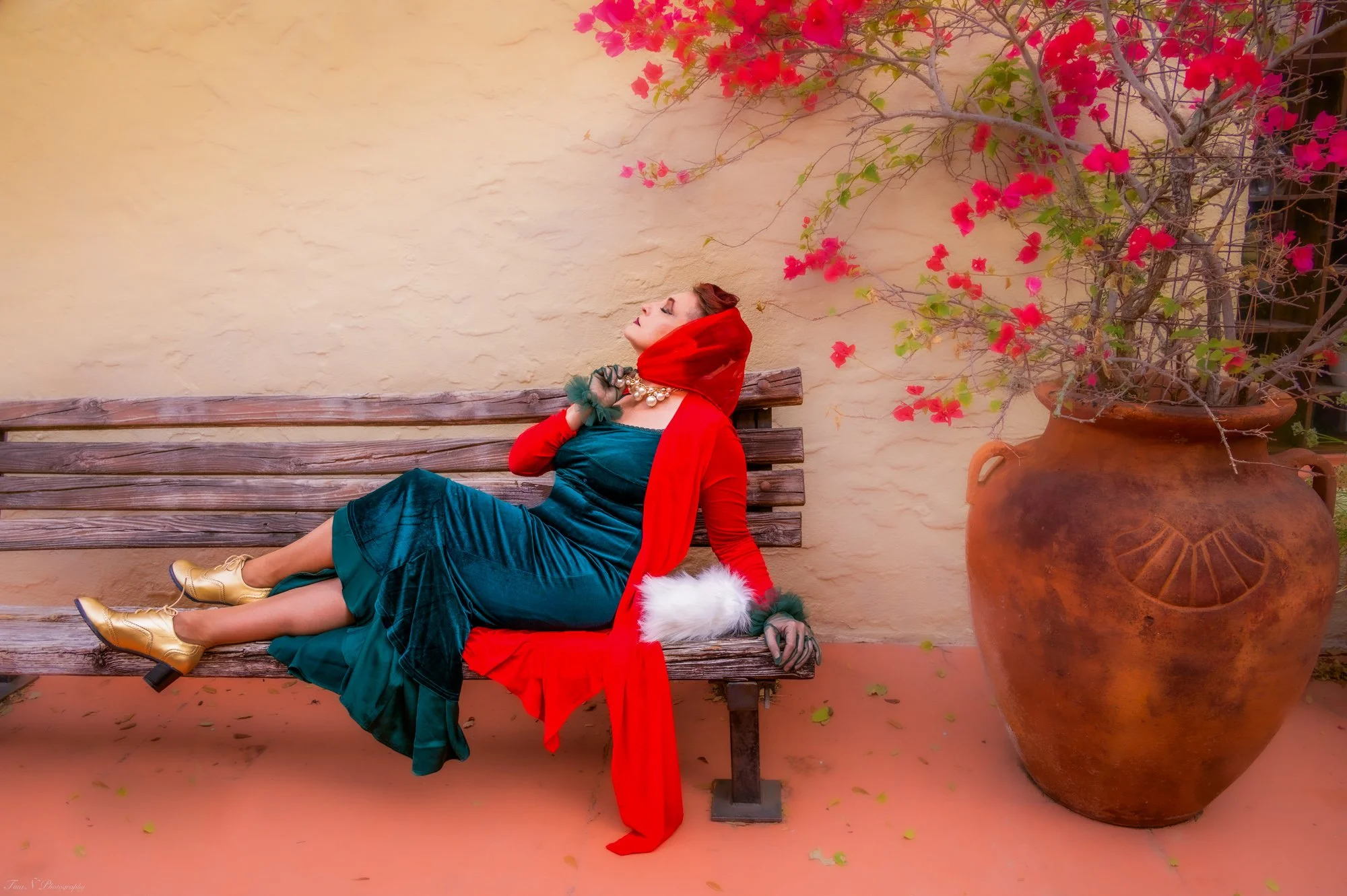 A woman dressed in vintage clothing, including a red hooded cape with white fur trim, a teal velvet dress, golden ankle boots, and a pearl necklace, is reclining on a wooden bench outdoors, with her eyes closed and head tilted back. To her left, ther