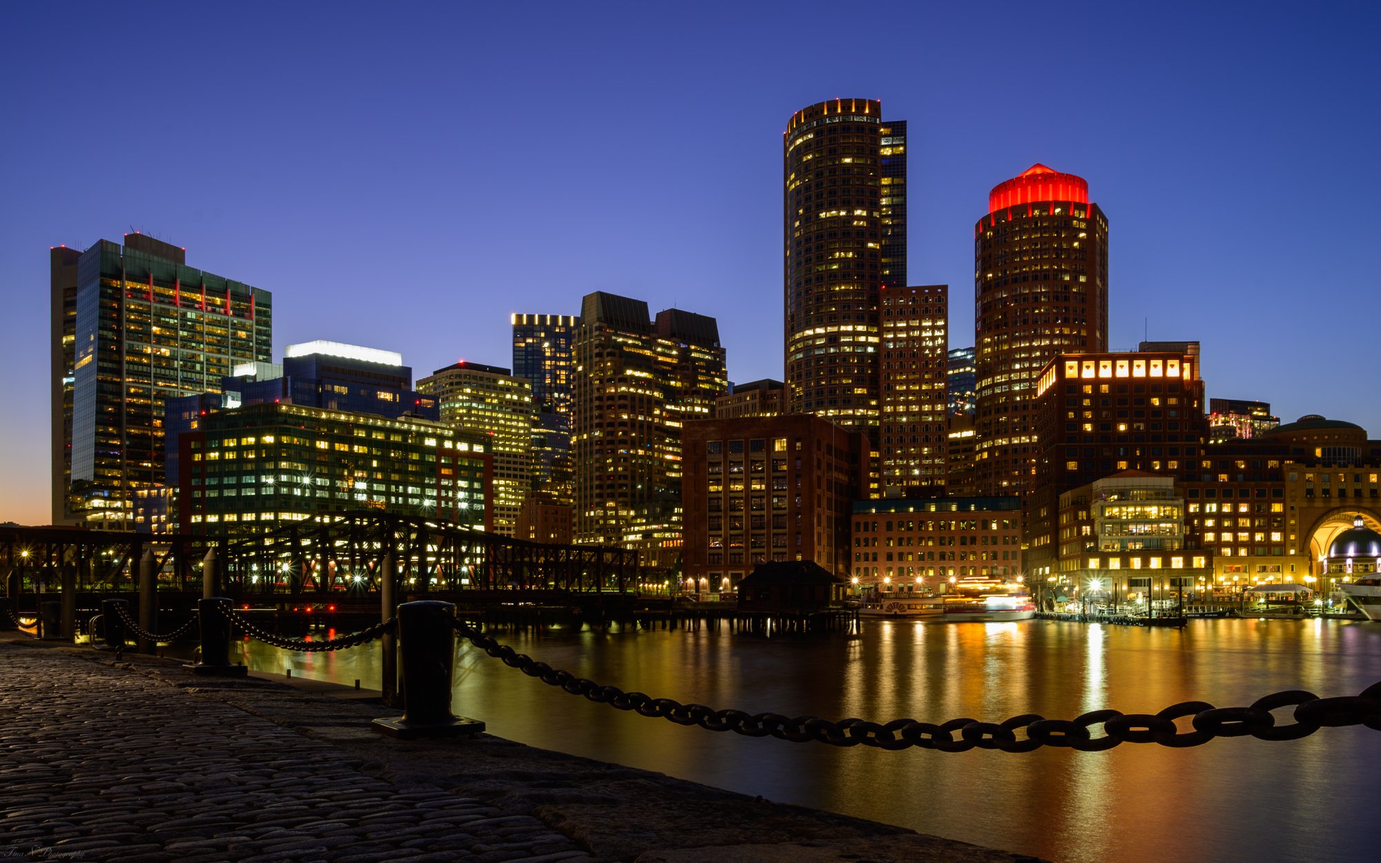 Night view of the Boston skyline with illuminated skyscrapers reflecting on the water, featuring a waterfront promenade with a chain barrier in the foreground. Boston city. 