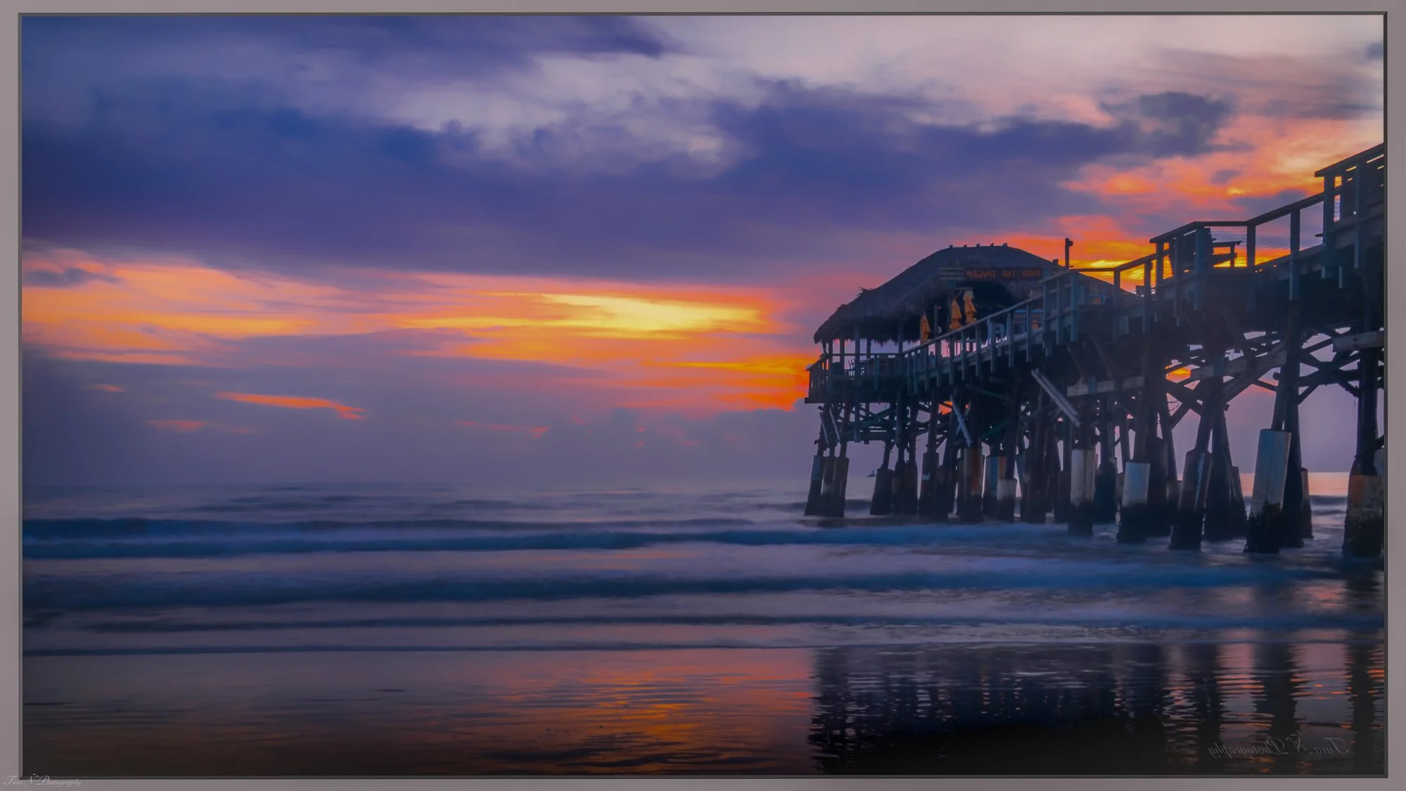 A wooden pier extends over the ocean during sunset, with purple and orange clouds in the sky and calm waves reflecting the colorful sky.