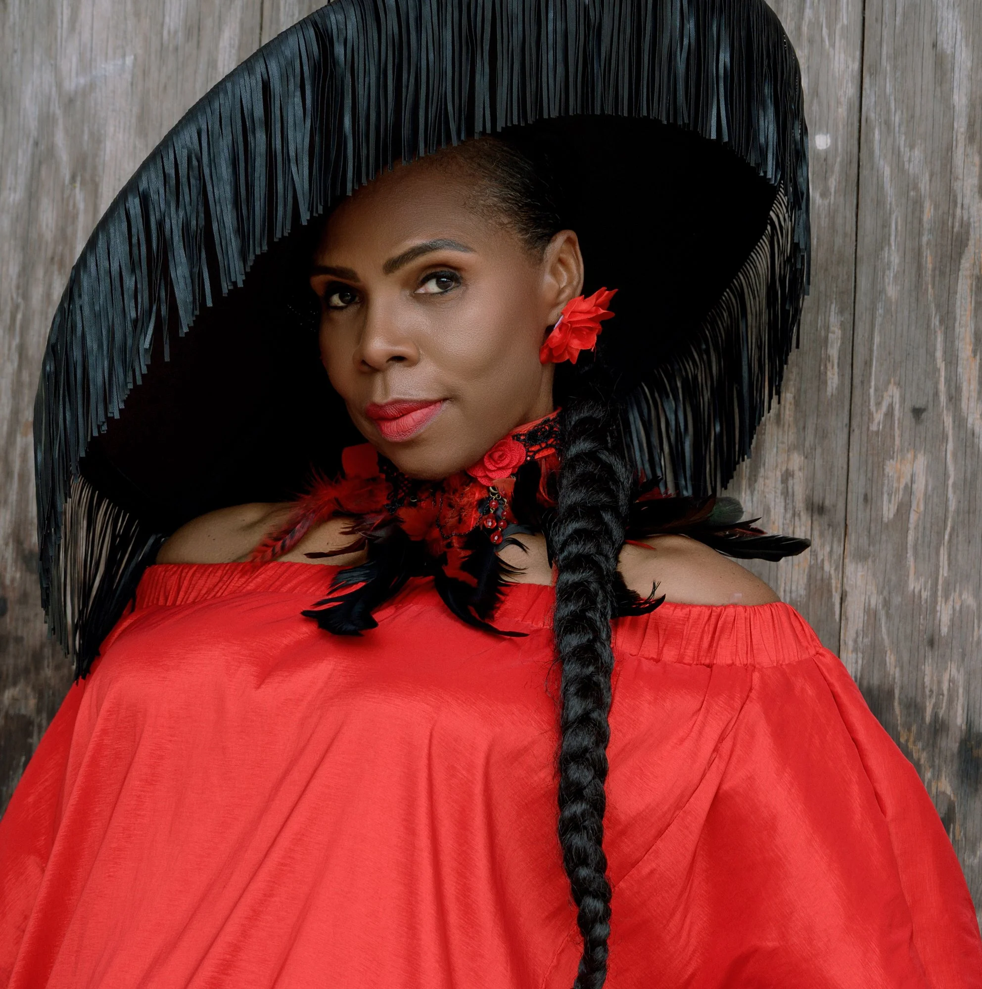 Woman with dark skin wearing a large black fringed hat, red dress, red flower earrings, and black feather details, standing against a wooden background.