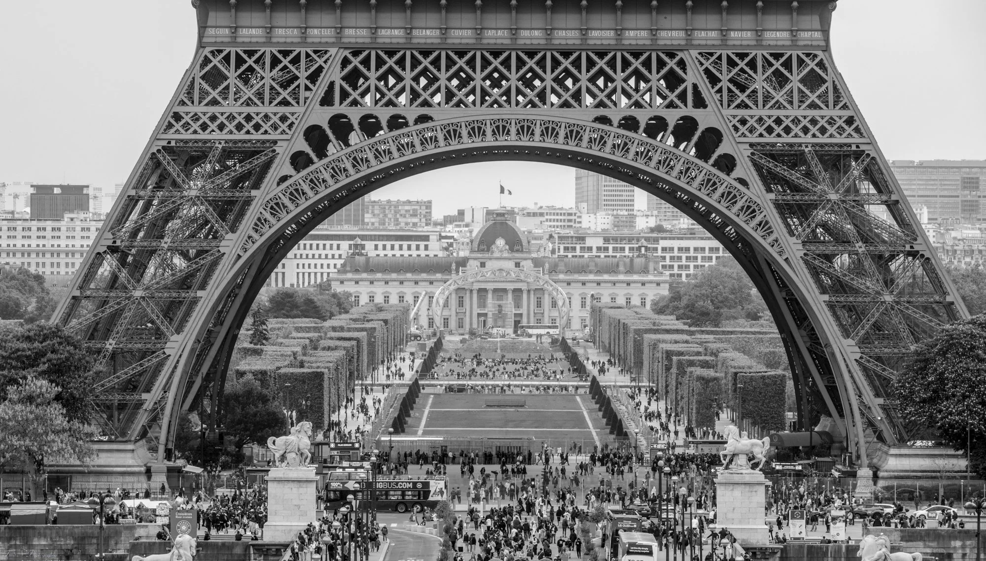 View of the Eiffel Tower from beneath, looking through its arch, with the Trocadéro Palace and gardens in the background, filled with visitors and trees.