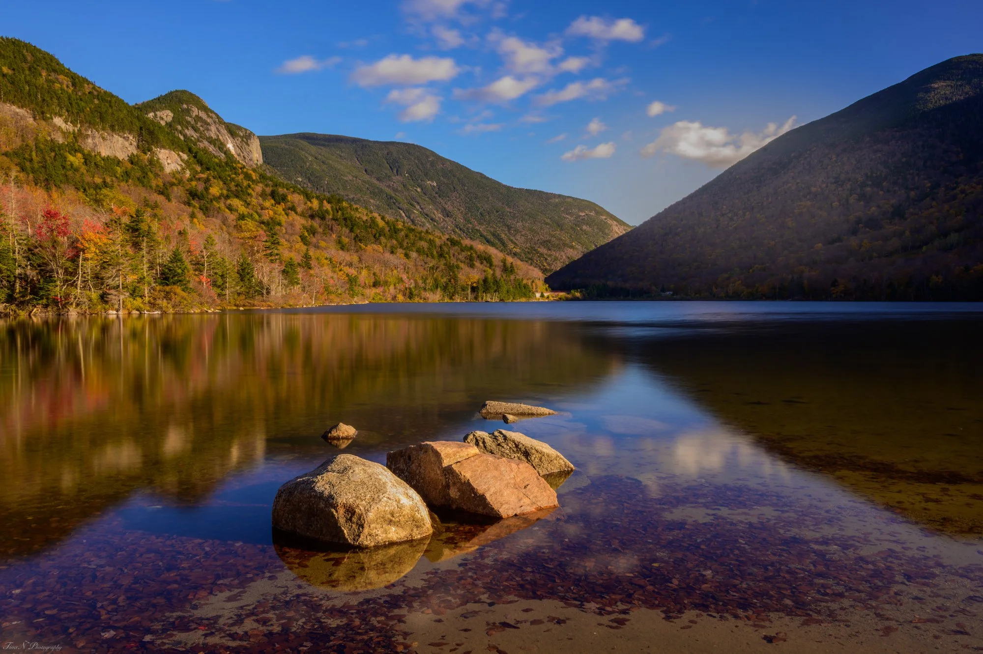 A mountain landscape with a calm river in the foreground, surrounded by lush green and fall-colored trees, under a bright blue sky with a few clouds.