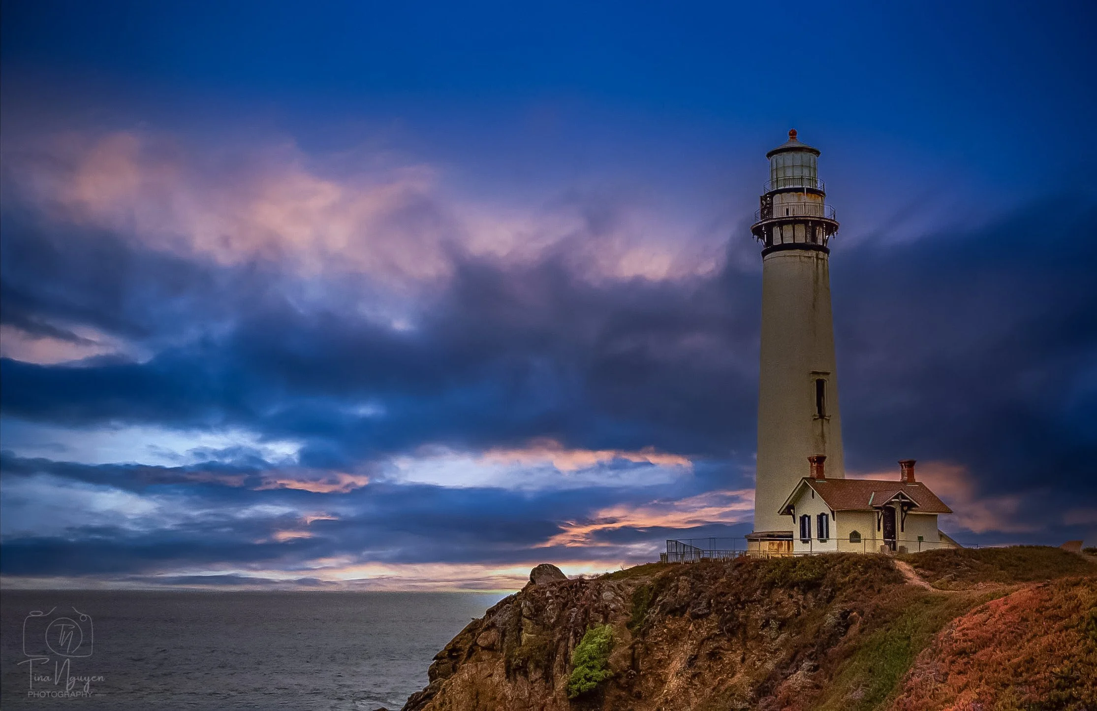 A lighthouse on a rocky cliff overlooking the ocean at sunset, with a building attached to it and cloudy sky.