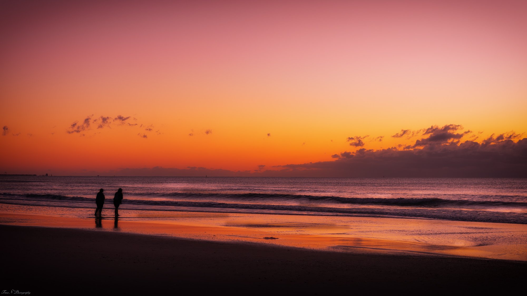 Silhouettes of two people walking along the shoreline at sunset with the sky painted in shades of pink, orange, and purple over the calm ocean. Cocoa Beach, Florida