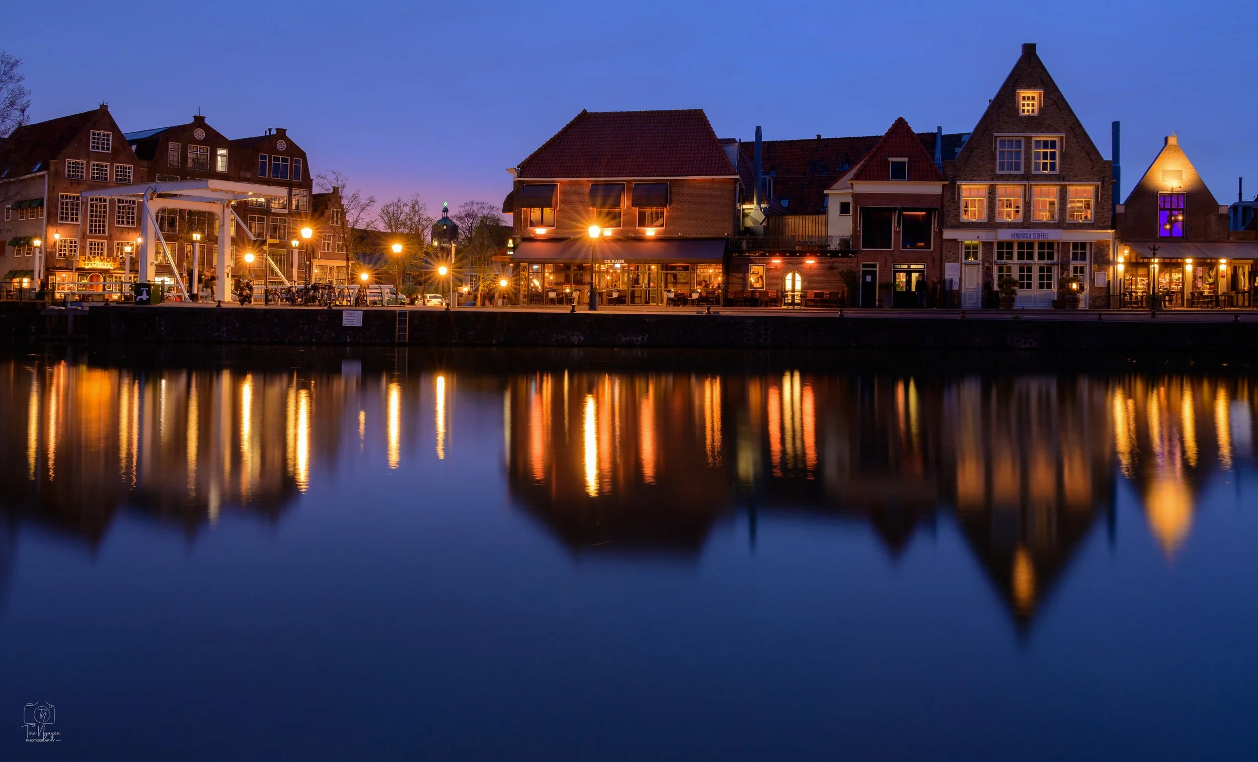 A serene night scene of illuminated buildings along a riverbank, reflecting warmly on the calm water.