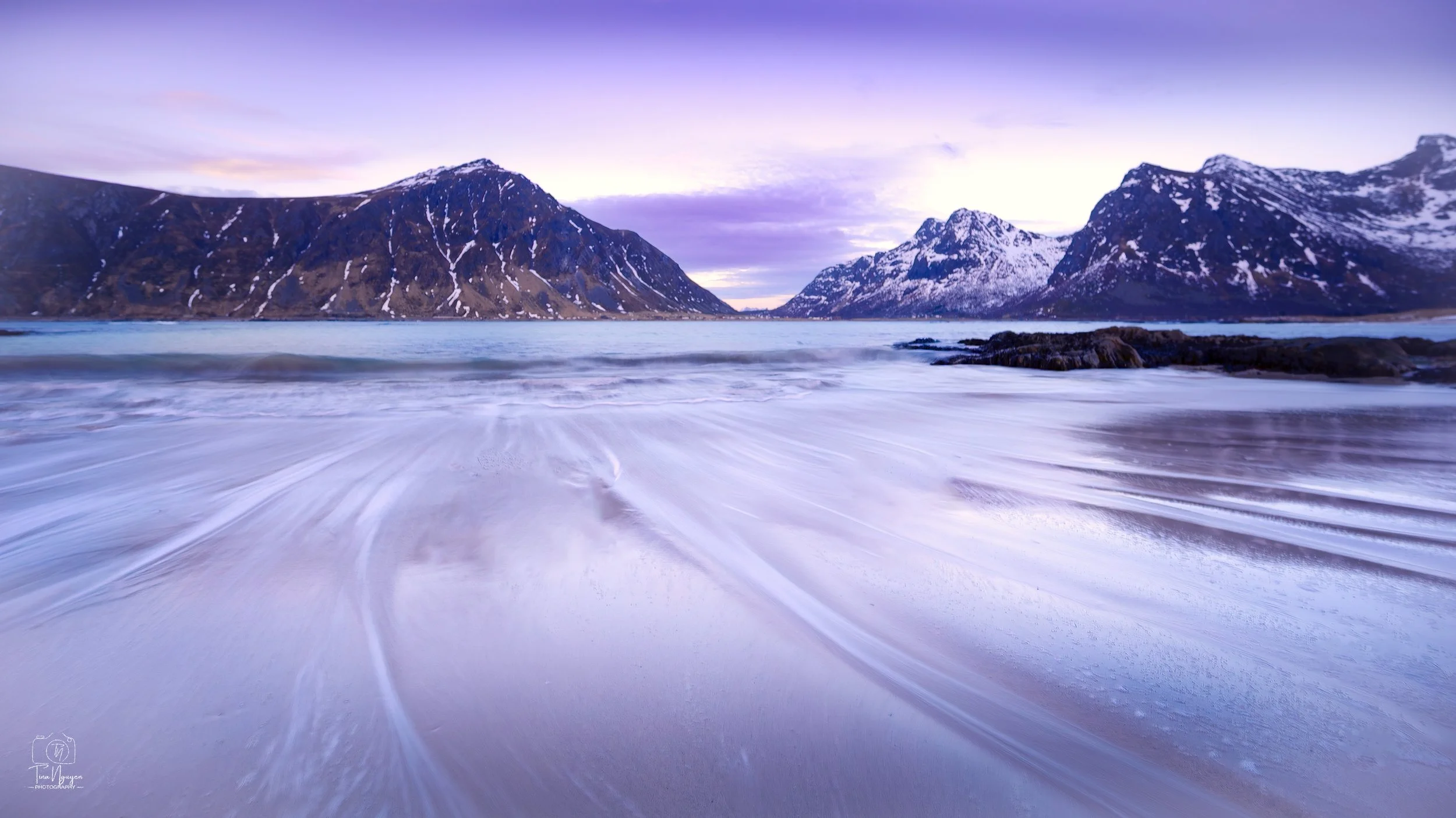Snowy mountains adjacent to a body of water with a sandy shore, under a cloudy sky