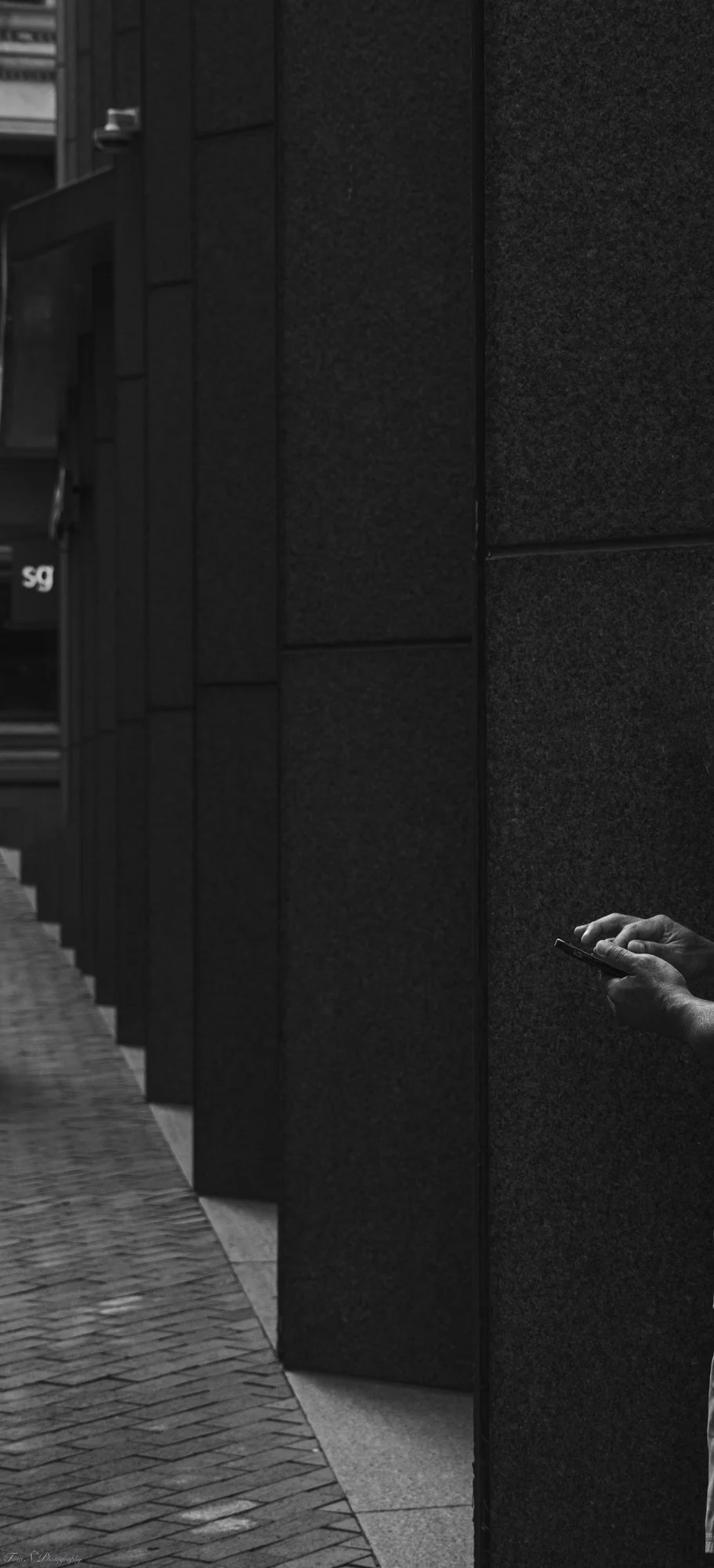 A person holding a smartphone with only their hands visible in a dark, urban setting next to a textured black wall, with a sidewalk on the left side of the image.