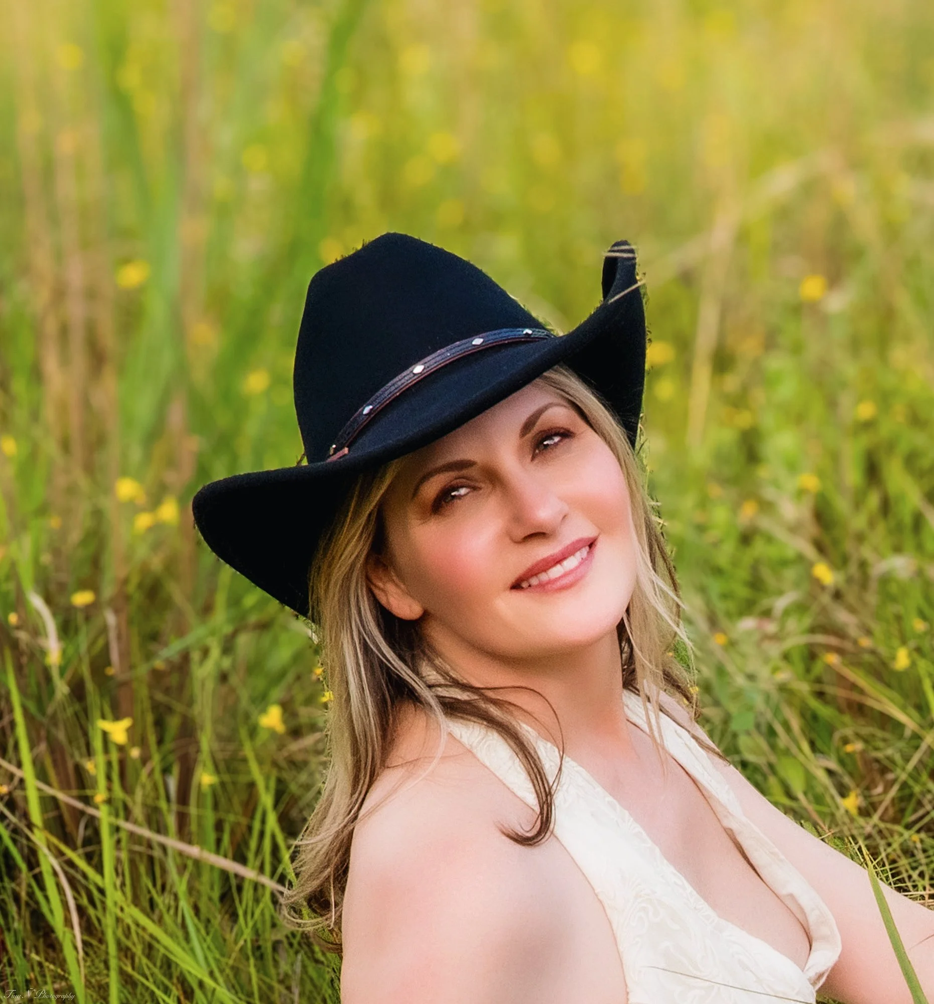 A woman with long hair wearing a black wide-brimmed hat and a white sleeveless top, smiling and looking up, surrounded by tall green grass and yellow flowers.