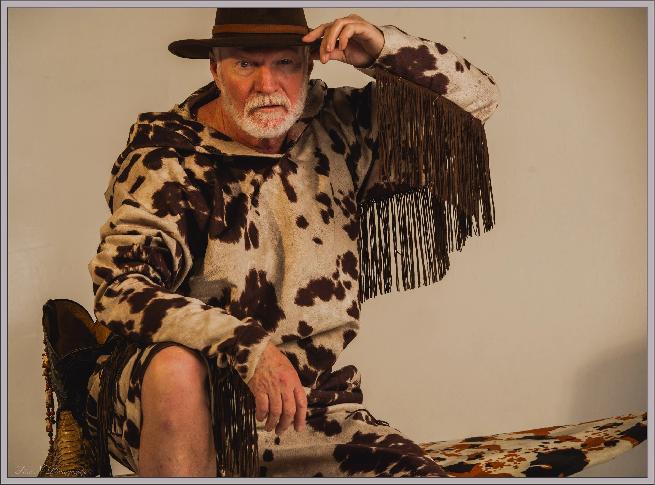 An older man dressed in a cow-patterned hoodie, cowboy hat, and carrying a cow-print ironing board cover, sitting on a stool with a cow-print sheep shearing tool to his right, posing with one hand on his knee and the other adjusting his hat.