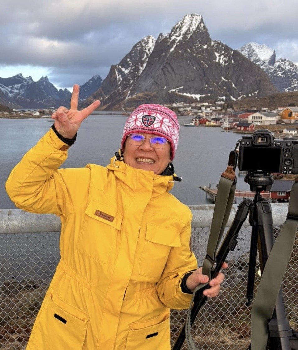 A woman in a yellow jacket and pink hat smiling and making a peace sign while holding a camera on a tripod, in front of a mountain landscape with snow-capped peaks, water, and small colorful buildings.