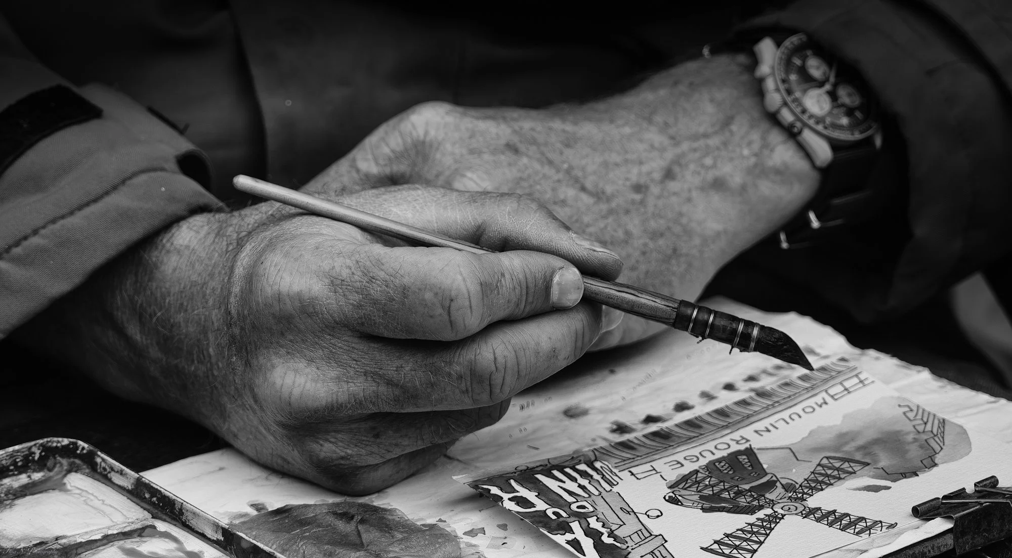 A black and white photo of an older person's hand holding a paintbrush, painting a colorful picture of a construction site with machinery and a crane on paper.