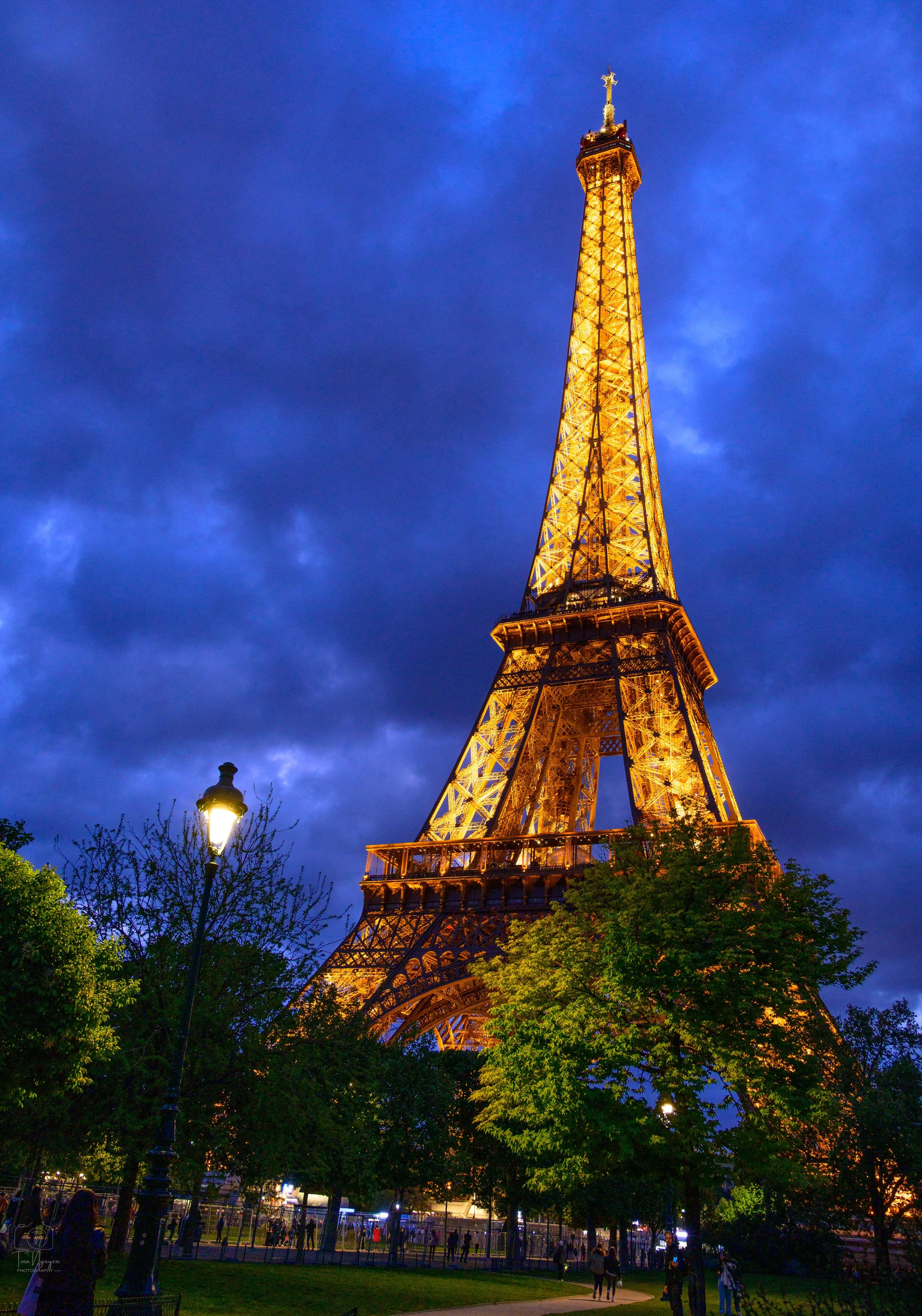 The Eiffel Tower illuminated at dusk with a cloudy sky in the background. There are trees, a lamppost, and people walking in the park at the base.