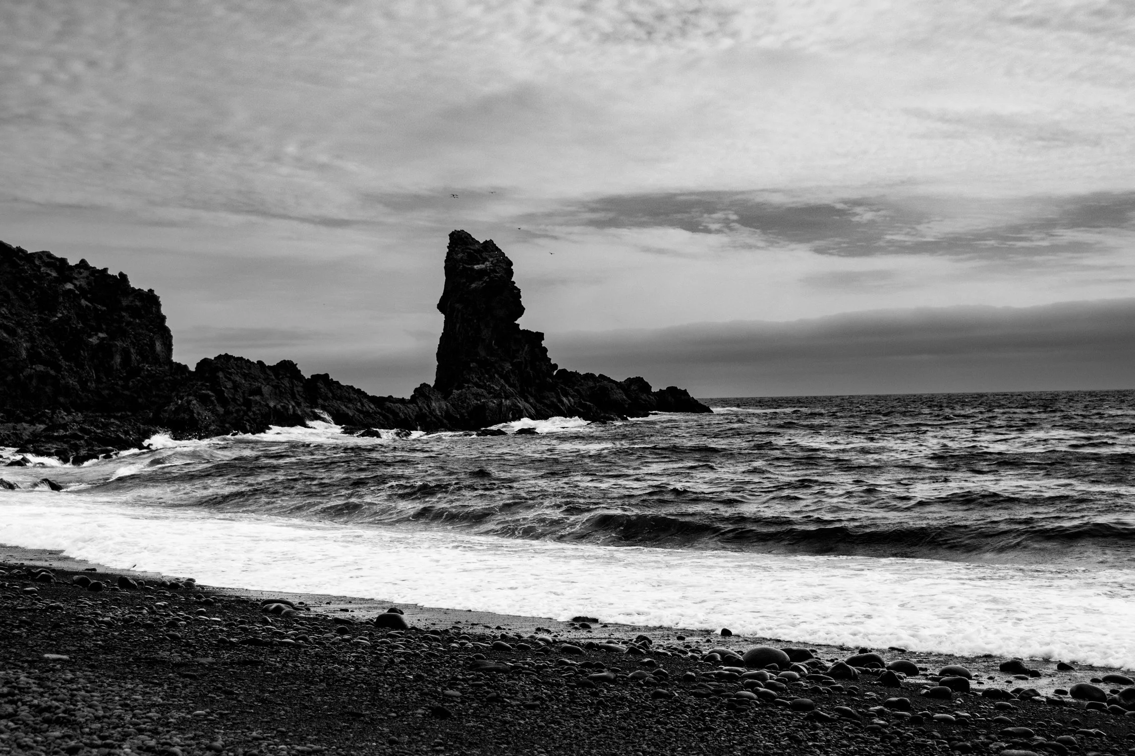 Black and white photo of a rocky coastline with a tall, jagged rock formation in the sea, pebble beach in the foreground, and cloudy sky overhead.