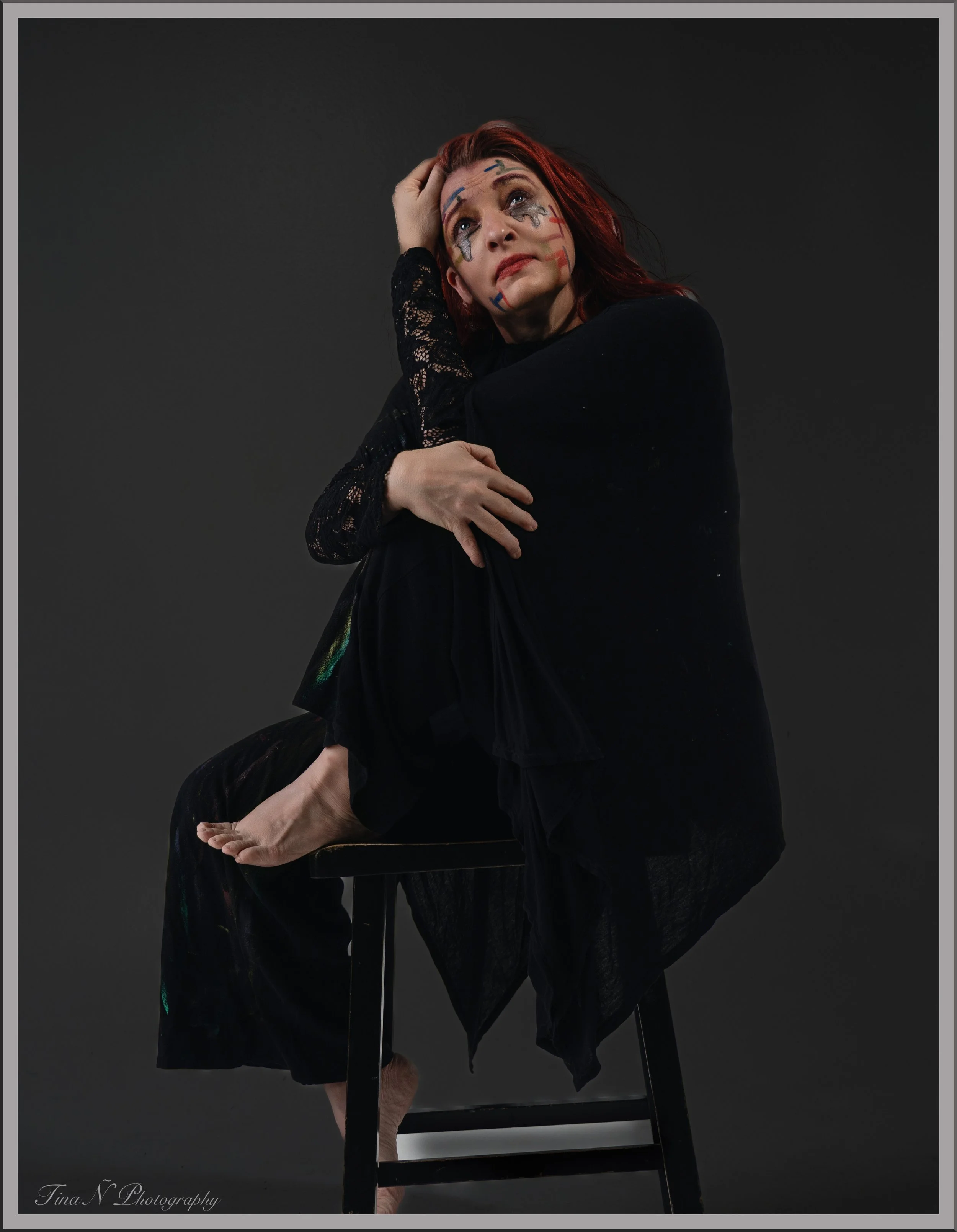 A woman with red hair, sitting on a black chair against a dark background, wearing a black lace dress, with painted facial markings, sitting with her legs crossed on the chair, looking upward with a contemplative expression.