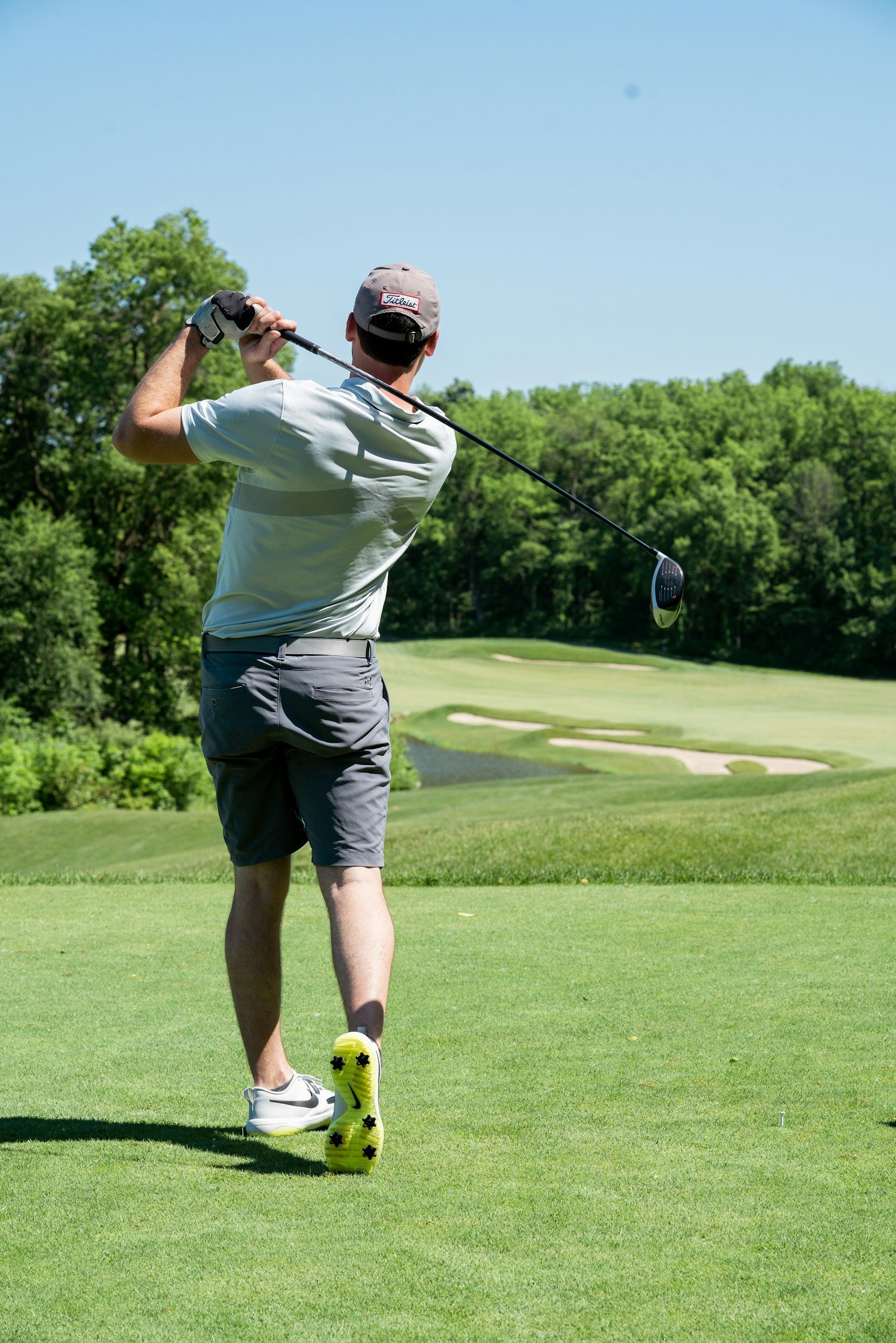 A man playing golf on a lush green course, wearing a gray cap, white shirt, and gray shorts, with yellow and black golf shoes, swinging a golf club.
