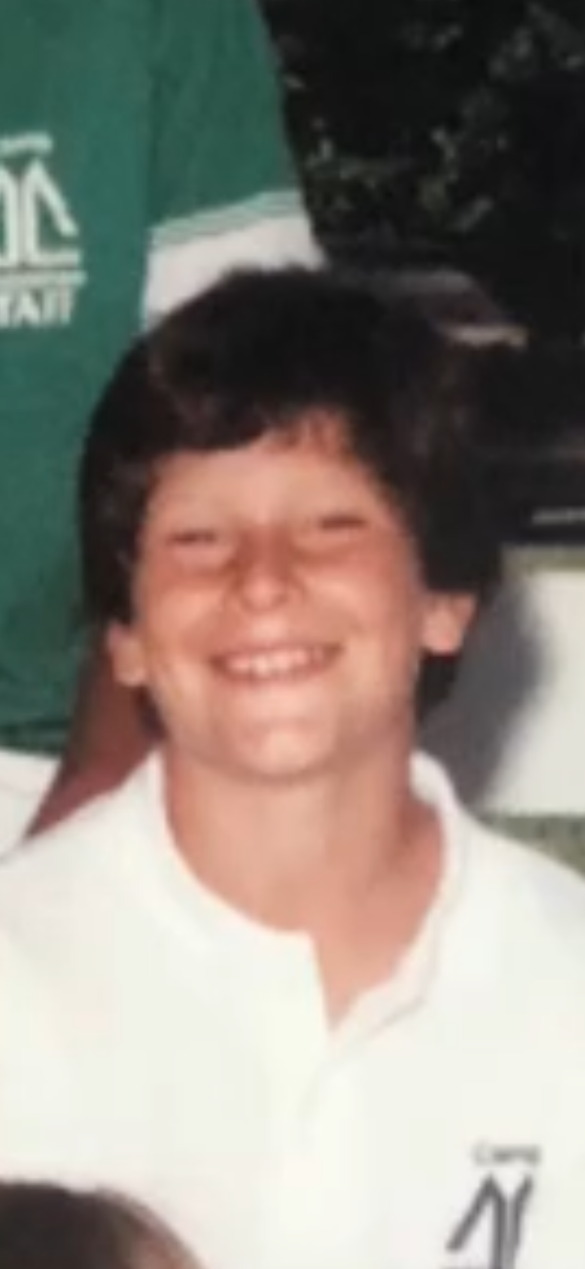 A smiling young boy with dark hair, wearing a white shirt, standing in front of a blurry outdoor background.