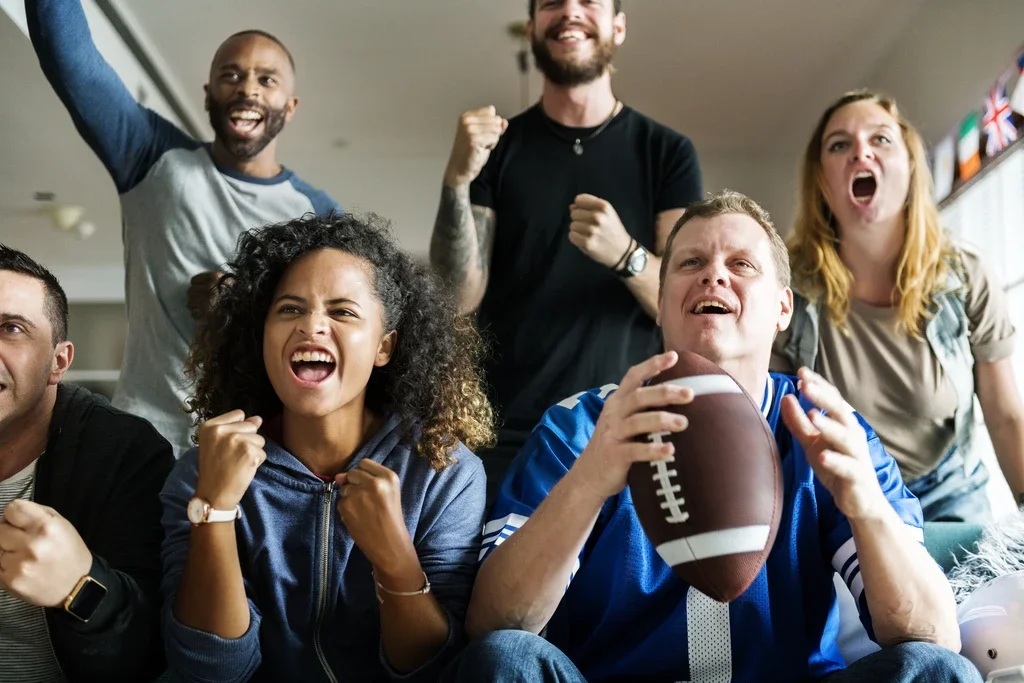 A group of six diverse people watching a football game and cheering enthusiastically.