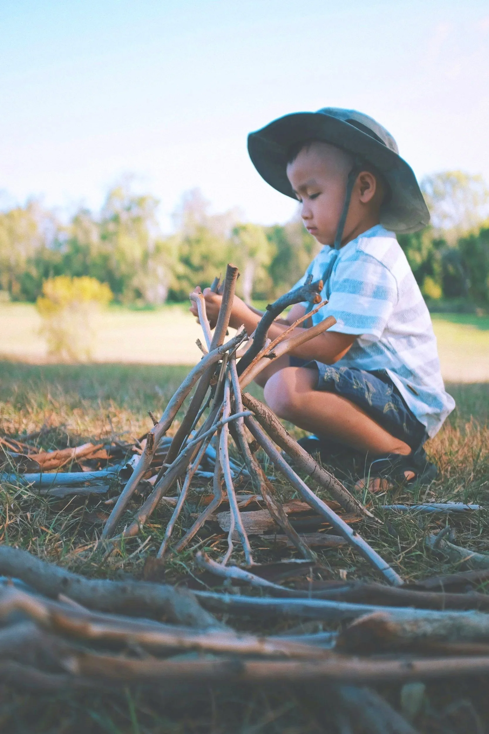 Young boy wearing a large hat, squatting outdoors and assembling a structure with sticks and branches.