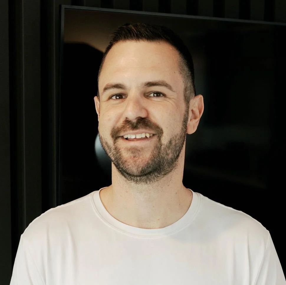A man with short dark hair and a beard, smiling, wearing a white t-shirt, standing in front of a dark background.