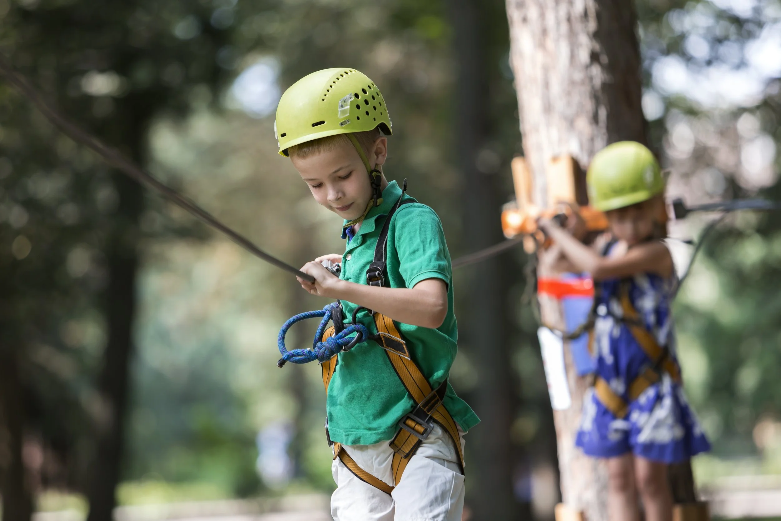 Children in safety harnesses and helmets rock climbing in a forested outdoor environment.