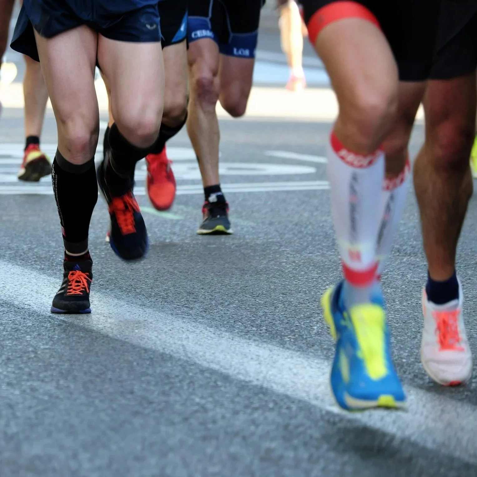 Close-up of multiple runners' legs and feet during a race, wearing athletic shoes and shorts, on a paved road.
