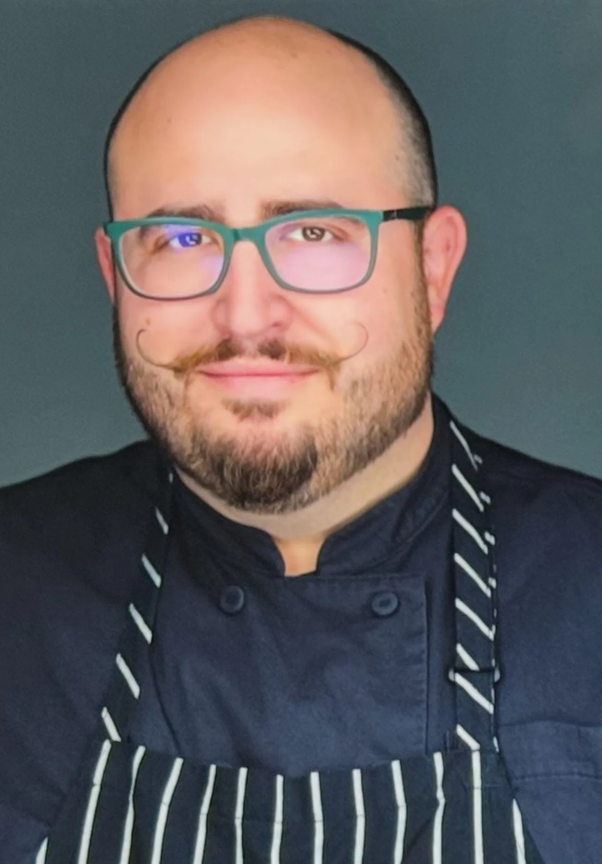 A man with glasses and a beard, wearing a black chef's jacket and a striped apron, smiling against a dark background.
