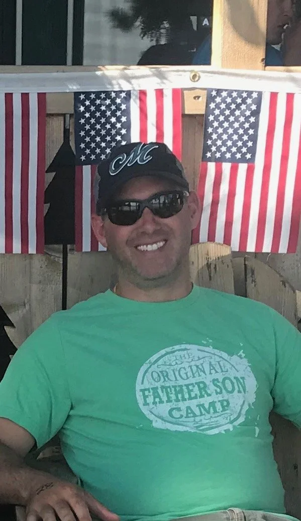 A man wearing sunglasses and a black cap with a cursive "M" is smiling while sitting outdoors. Behind him are American flags displayed on a wooden fence. He is wearing a green T-shirt that reads "The Original Father Son Camp."