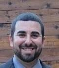 Close-up photo of a man with dark hair and a beard, smiling, wearing a suit jacket and shirt, against a wooden background.