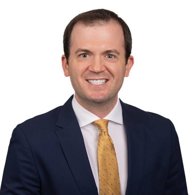 A smiling man in a navy suit, white shirt, and yellow tie against a white background.