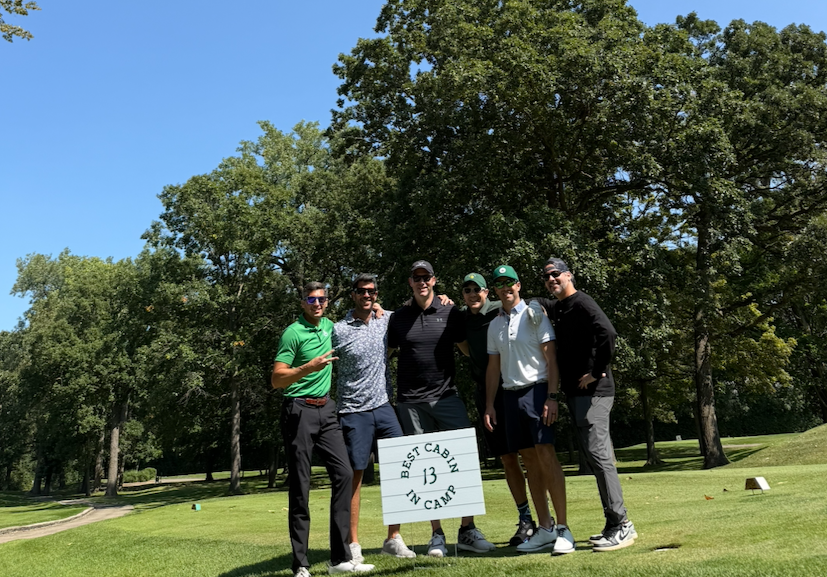 Group of six men standing on a golf course holding a sign that says "Best Cab in Camp" with the number 13, surrounded by trees and a clear blue sky.