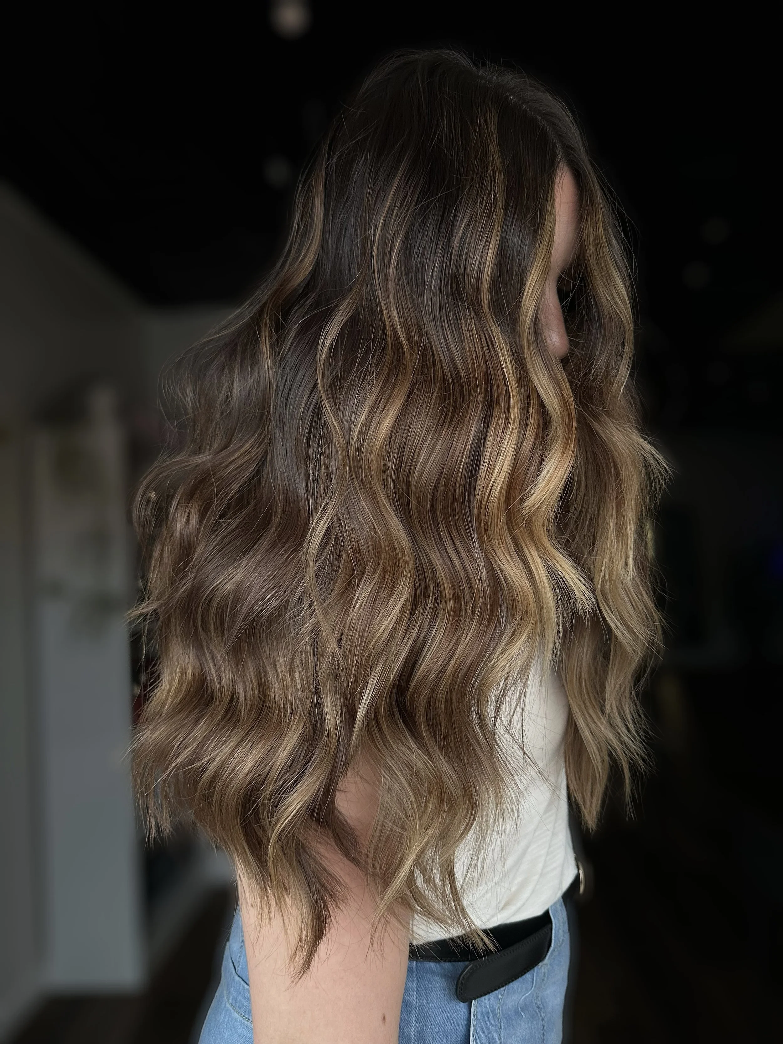 Side view of a woman with long, wavy, ombre hair transitioning from dark brown at the roots to blonde tips, wearing a white top and light blue jeans.