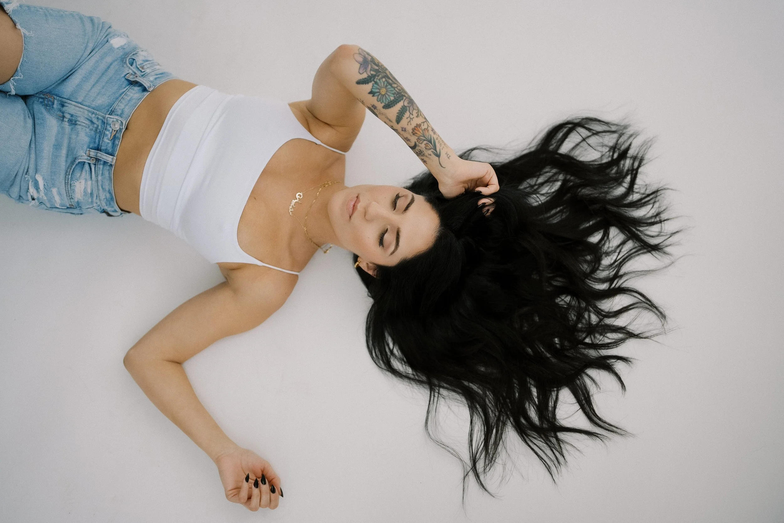 A woman with long black hair, lying on a white surface, wearing a white tank top and denim shorts, with floral tattoos on her arm, posing with her eyes closed.