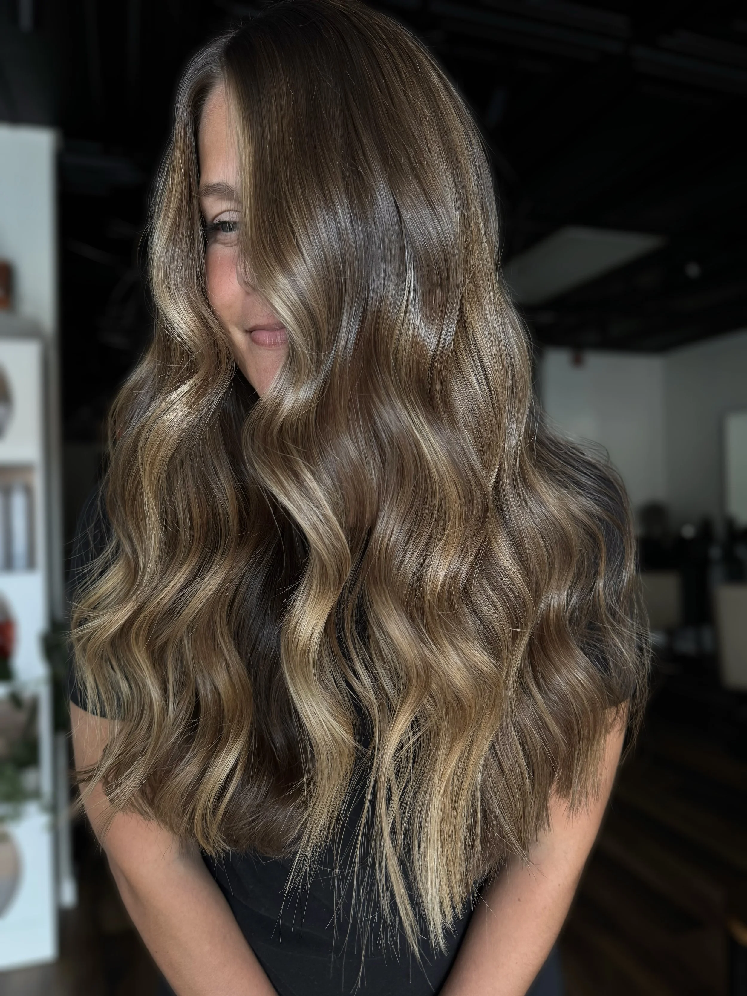 A woman with long, wavy, light brown hair with highlights, smiling softly and looking down in a salon setting.