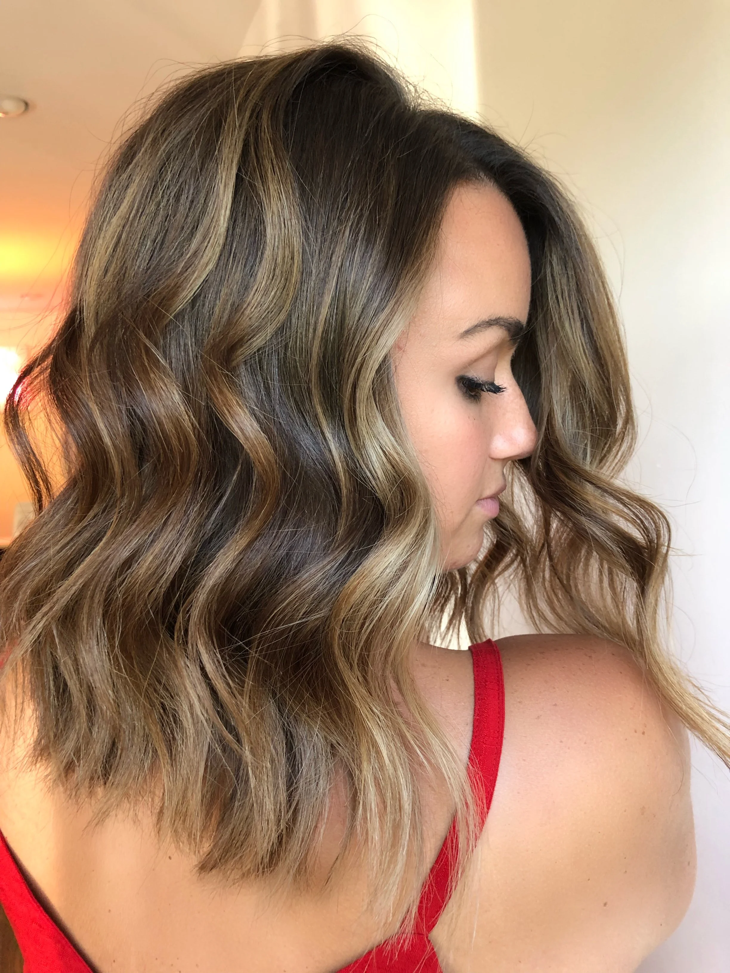 A woman with shoulder-length wavy hair with highlights, wearing a red top, looking down with her eyes closed.