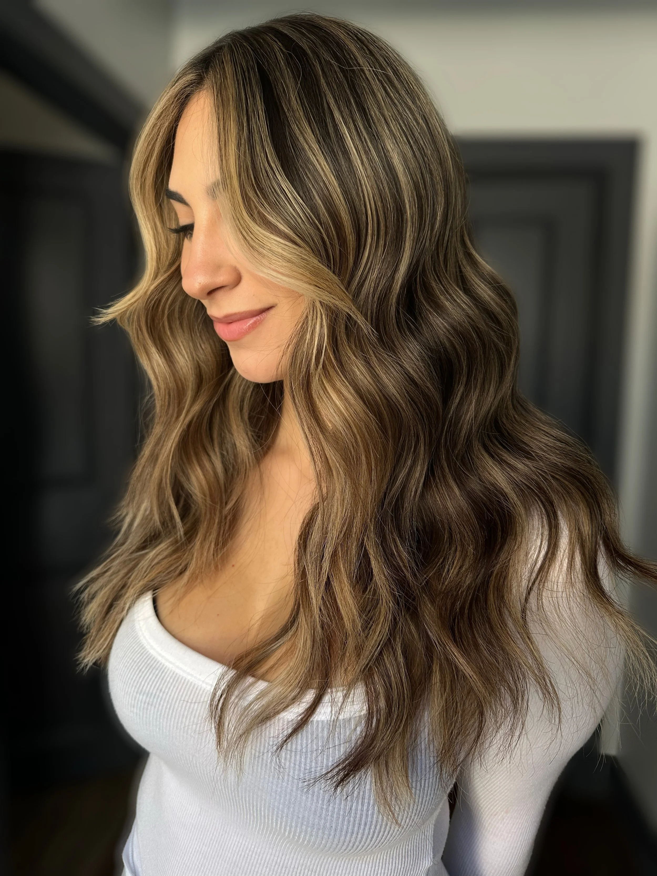 A woman with long, wavy, highlighted brown hair wearing a white top, looking down with a gentle smile.