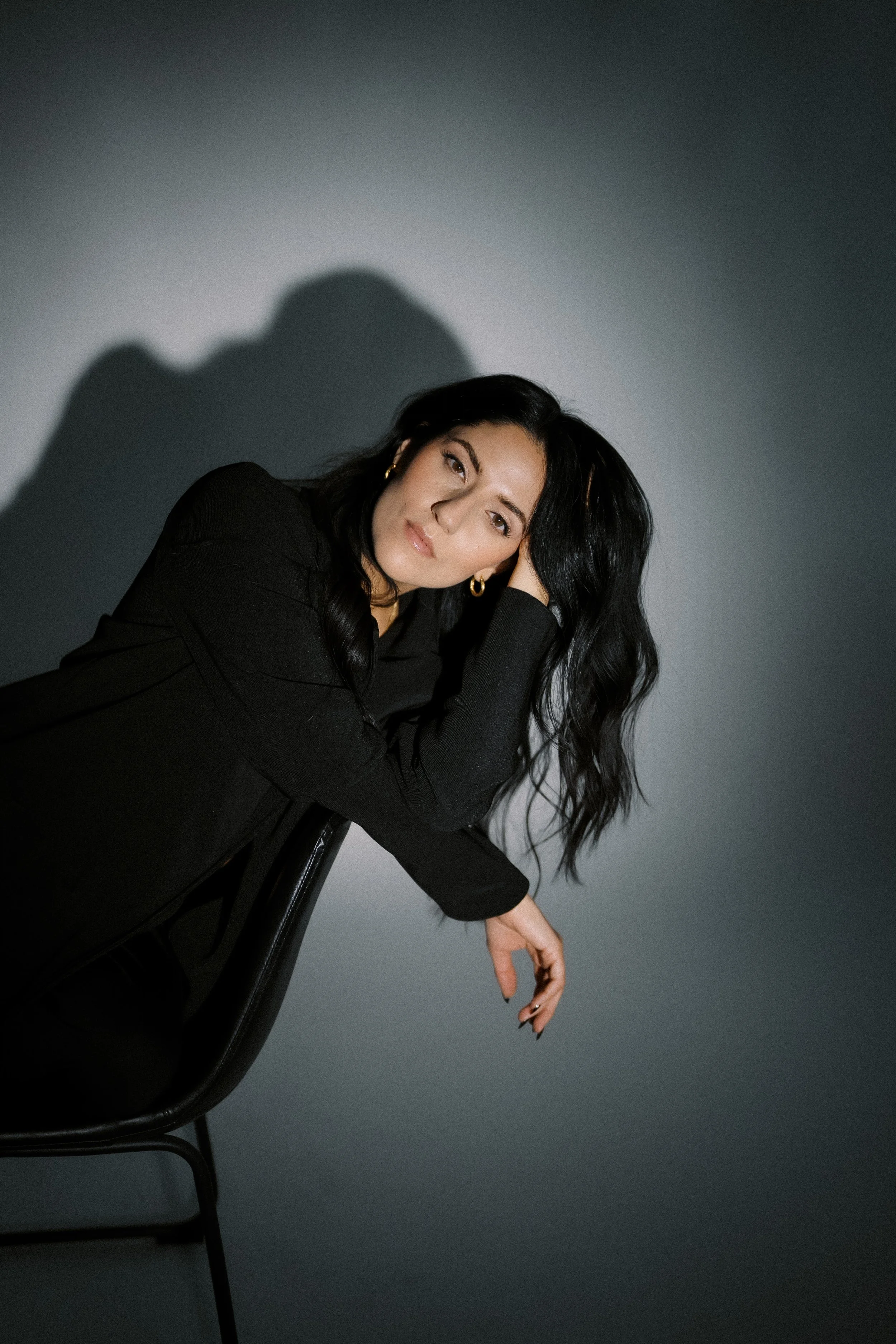 A woman with long black hair, wearing a black outfit and gold earrings, poses leaning on a black chair against a gray background with a shadow cast on the wall.