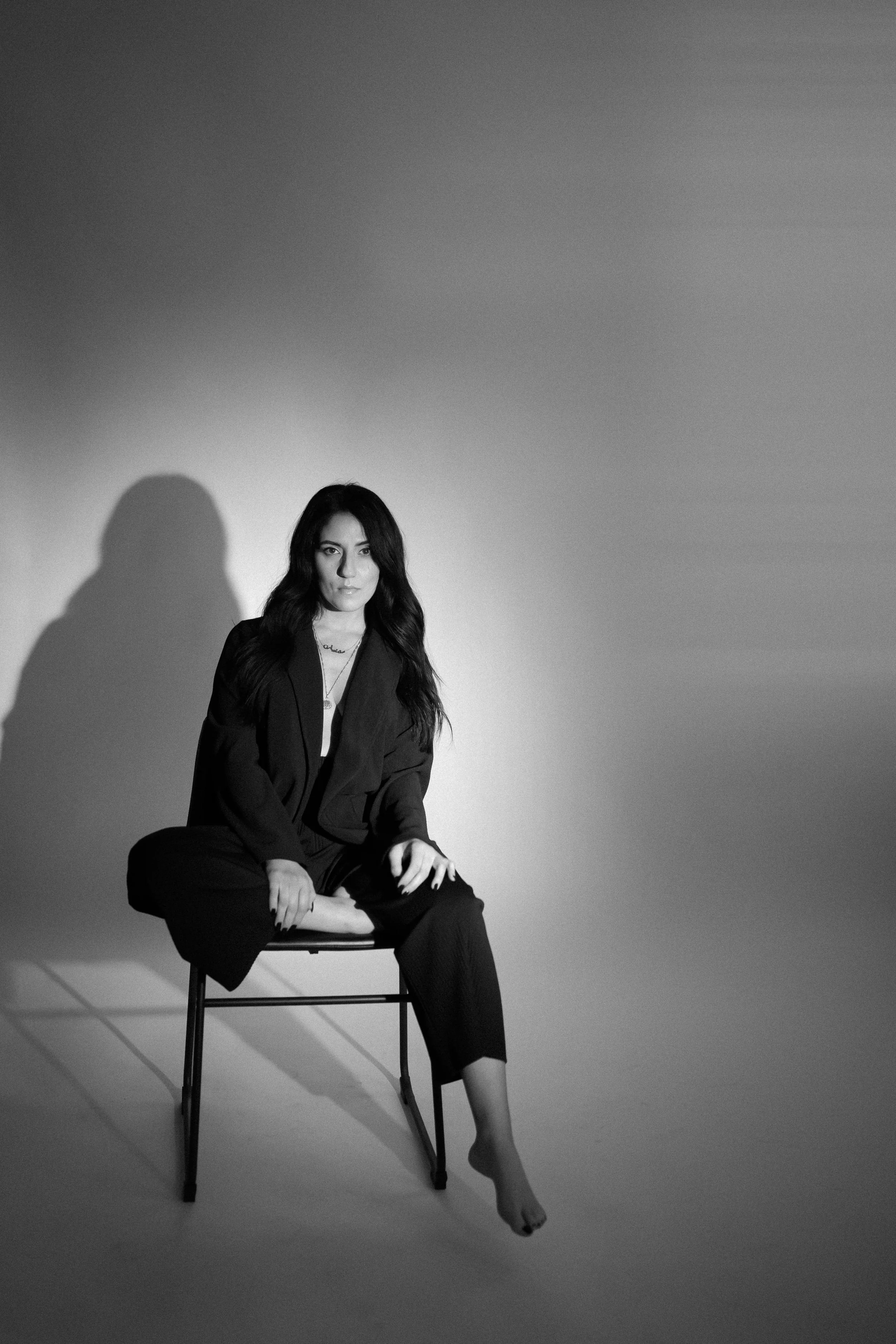 A woman with long dark hair sitting on a chair in a black suit, with one foot on the ground and the other leg crossed on her lap, against a plain background with shadows.