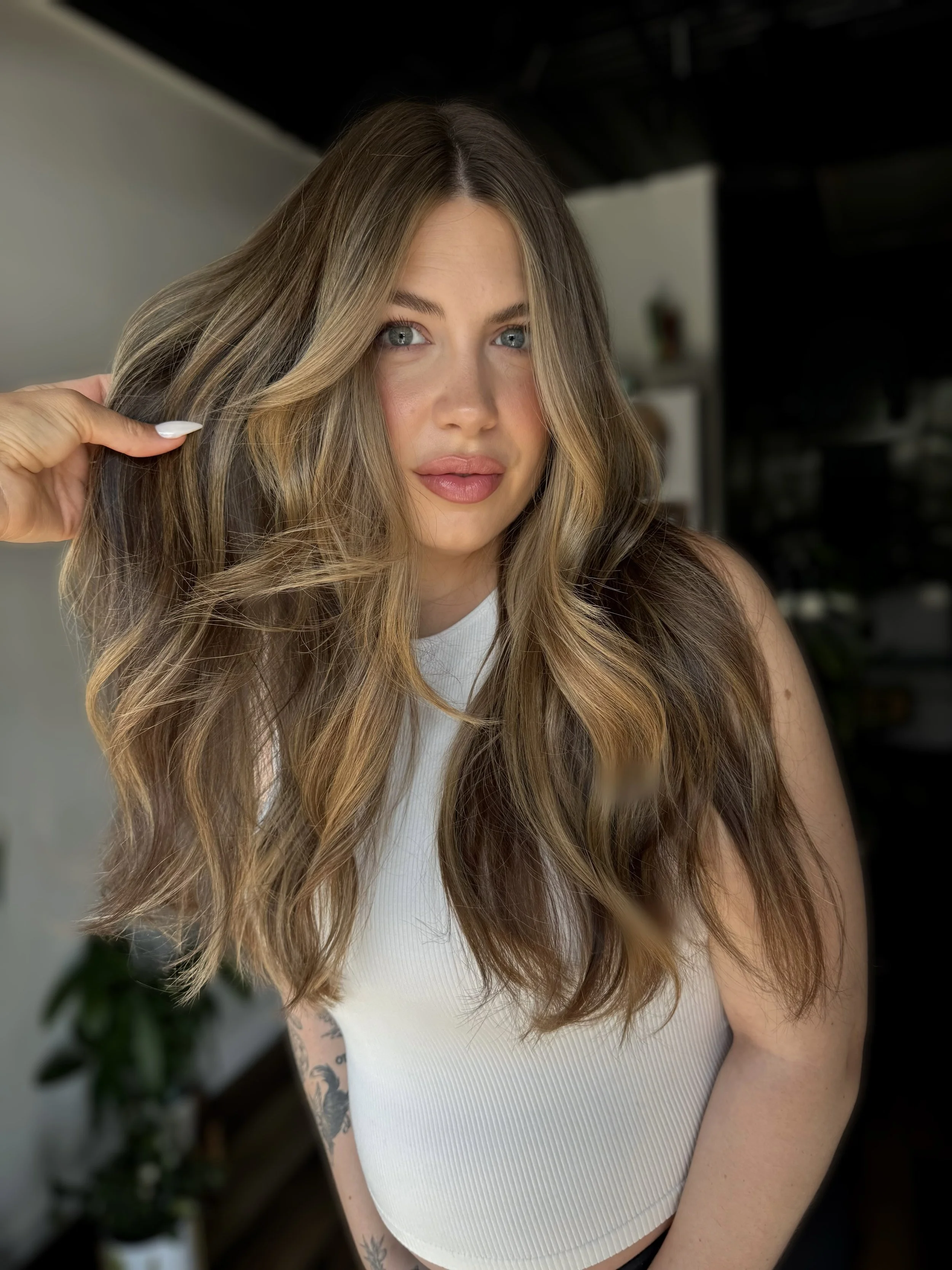 A young woman with long wavy hair, blue eyes, and light makeup, wearing a white sleeveless top, posing indoors with a hand touching her hair, in a well-lit space with blurred background.