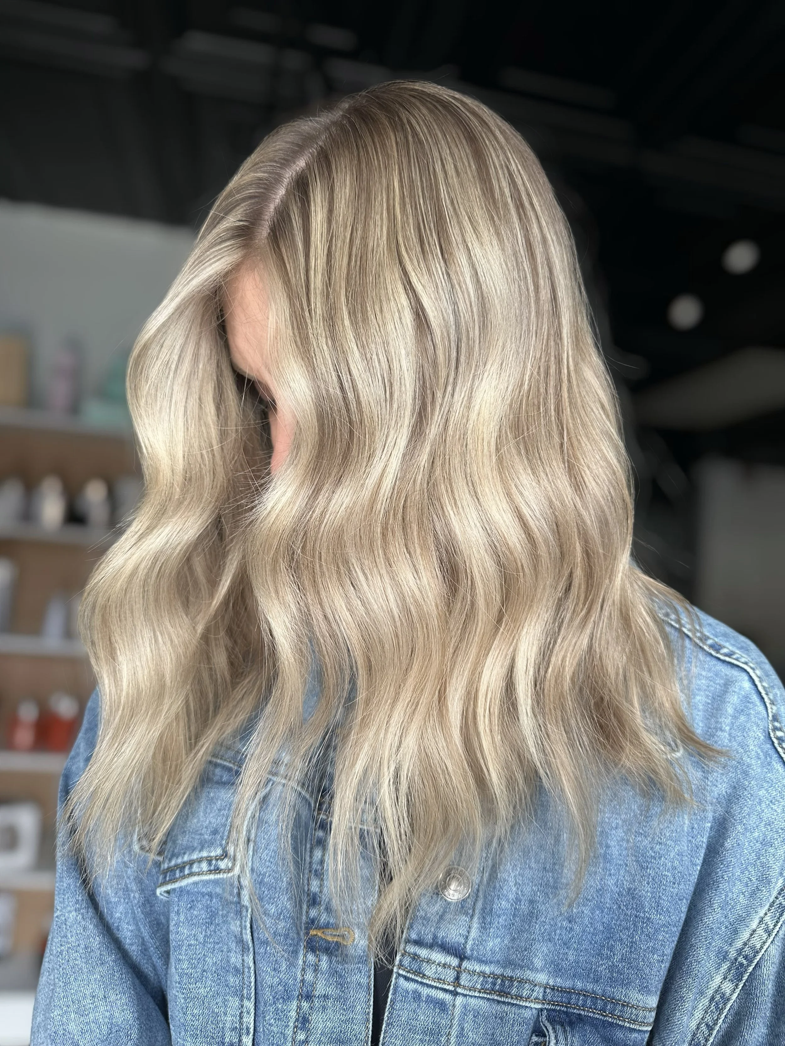 Close-up of a woman with long, wavy blonde hair wearing a blue denim jacket, indoors with shelves in the background.