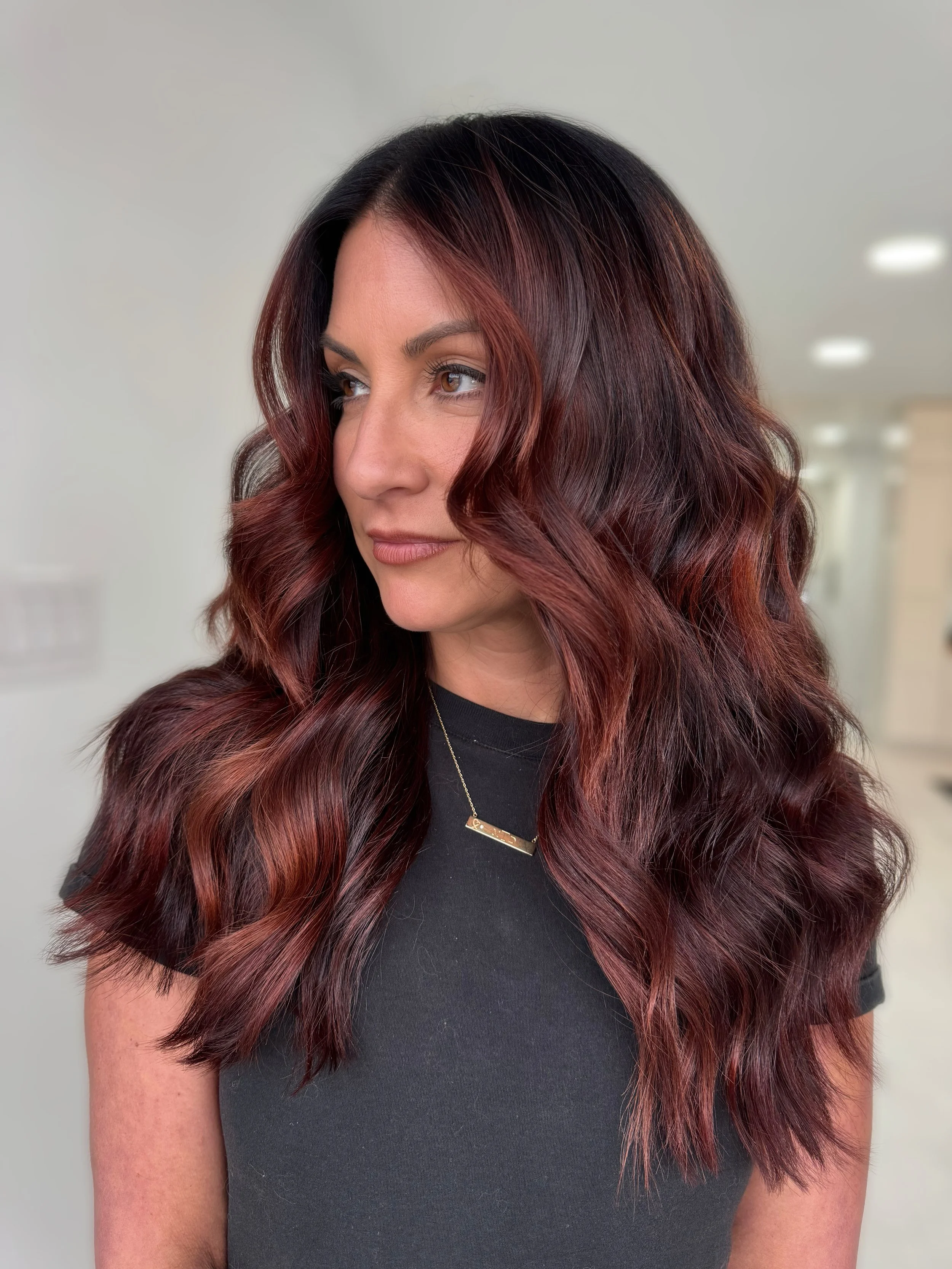 A woman with long, wavy dark brown hair with red highlights, wearing a black shirt and a gold necklace, looking to her left inside a bright, modern indoor space.