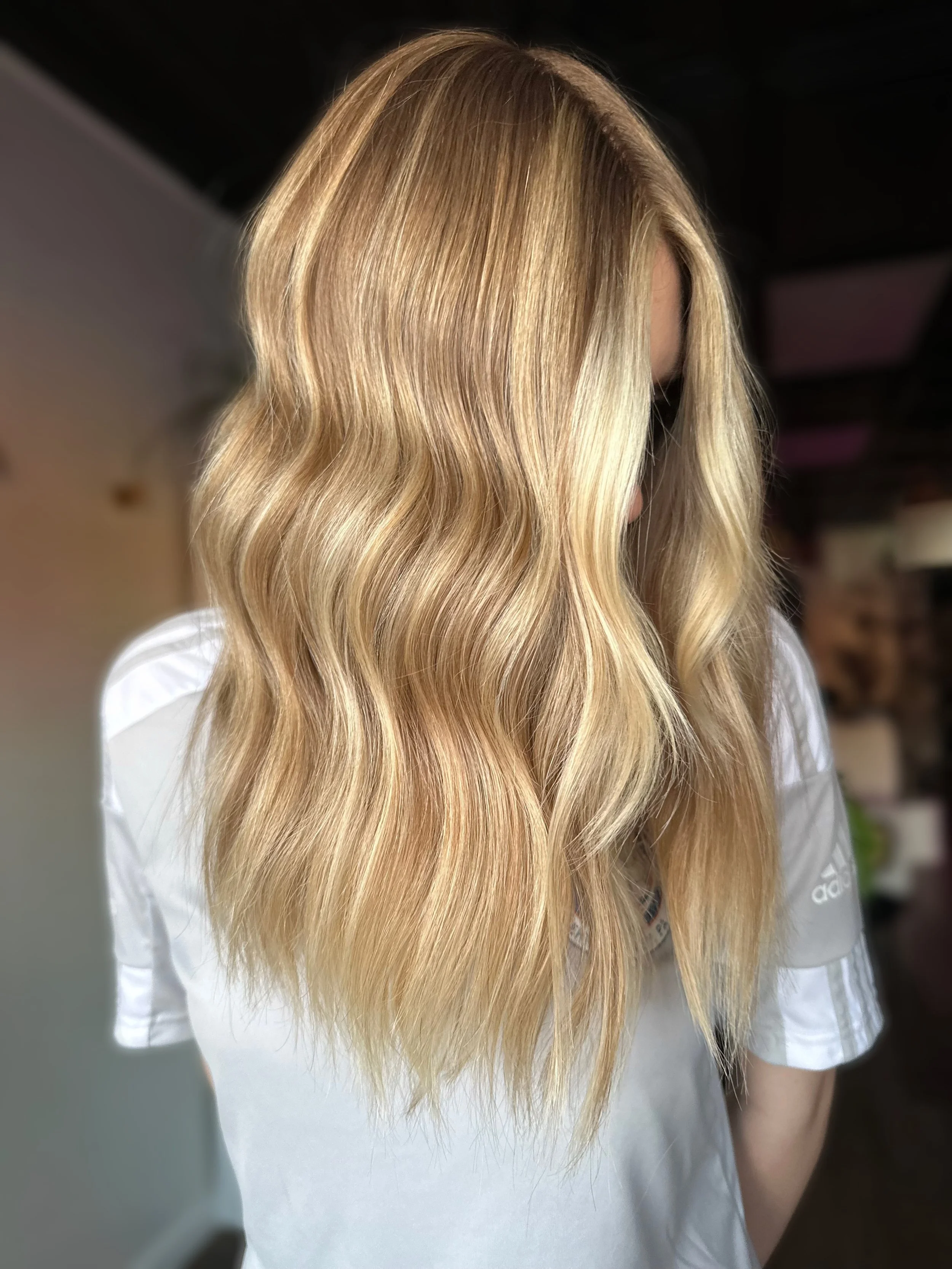 Close-up of a woman with long, wavy blonde hair, wearing a white shirt, standing indoors.