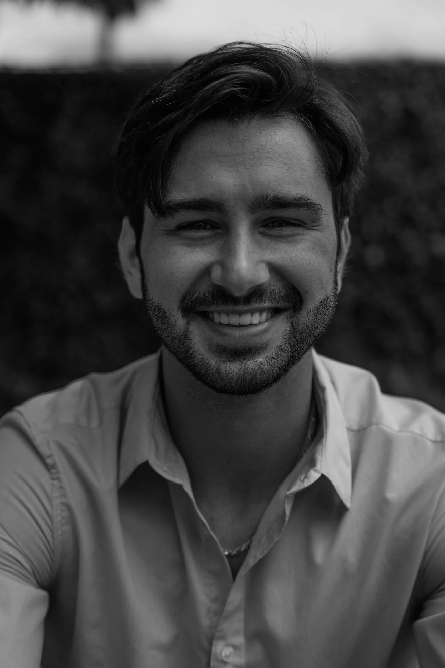 Portrait of a smiling young man with a beard, wearing a collared shirt, in black and white.