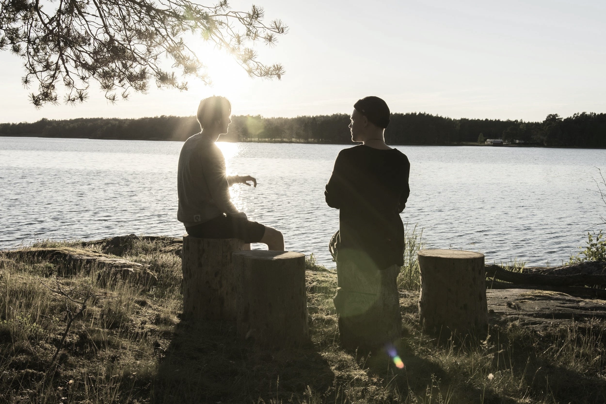 Two people sitting on tree trunks talking by a lake at sunset with sunlight glare and trees in the background.