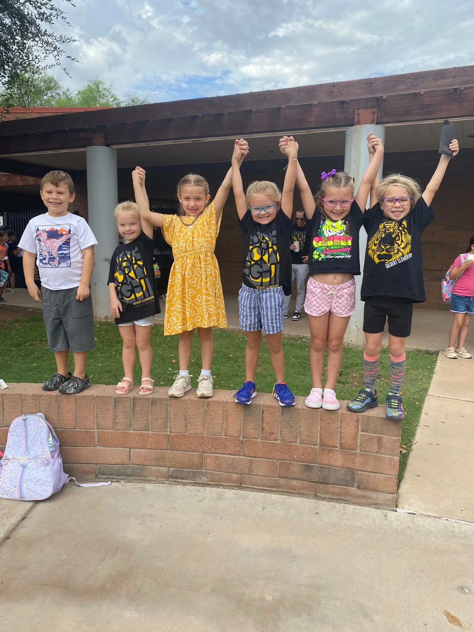 Six children standing on a brick ledge outdoors, holding hands and raising their arms in celebration. A backpack is on the ground in front of them, and a building with a porch and a cloudy sky are in the background.